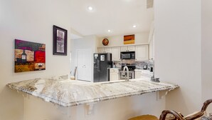 Uma cozinha com bancada de mármore, armários brancos, geladeira, micro-ondas e um relógio na parede.