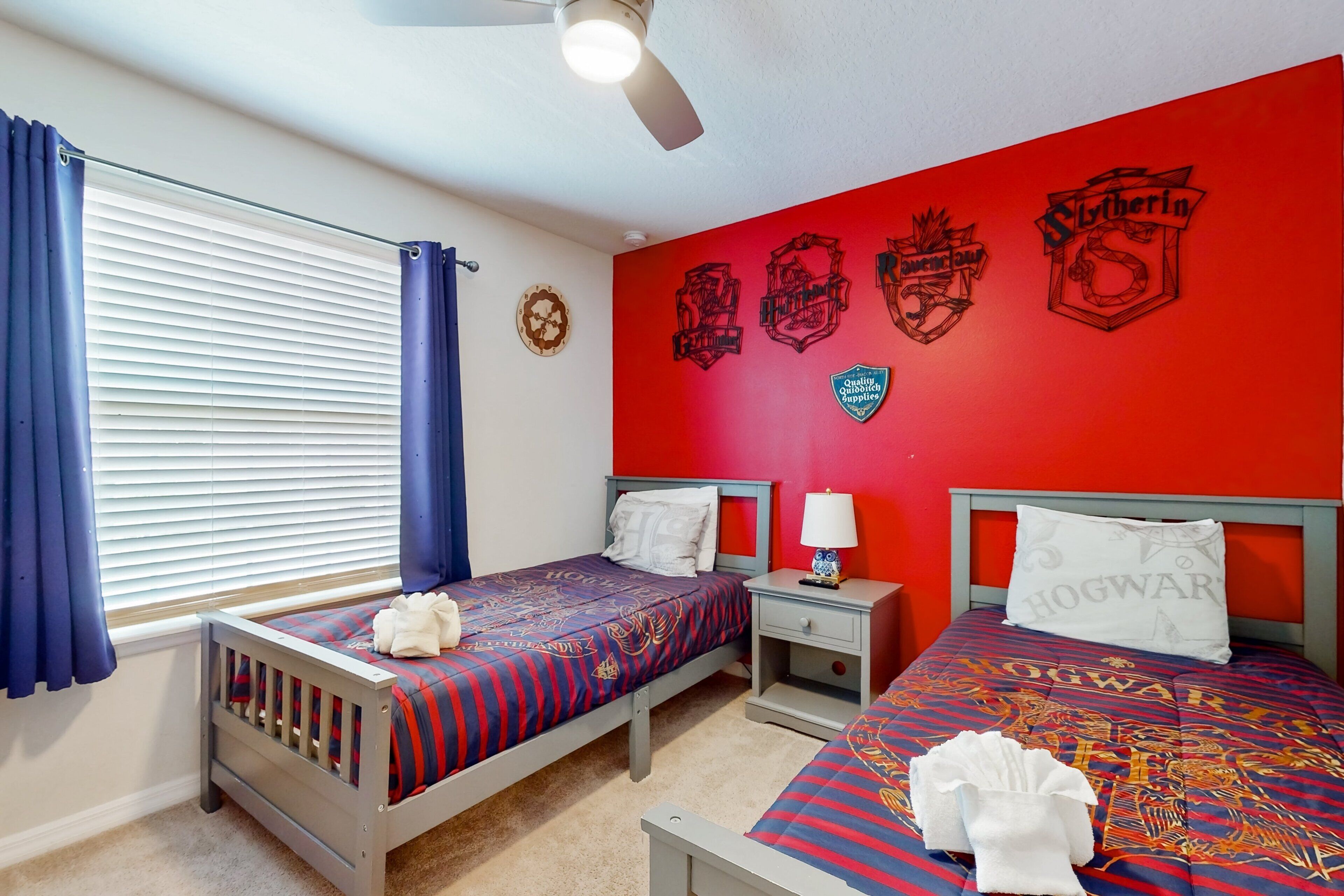 A bedroom with two beds, a window with blinds, a ceiling fan, and a red wall with various decorations.