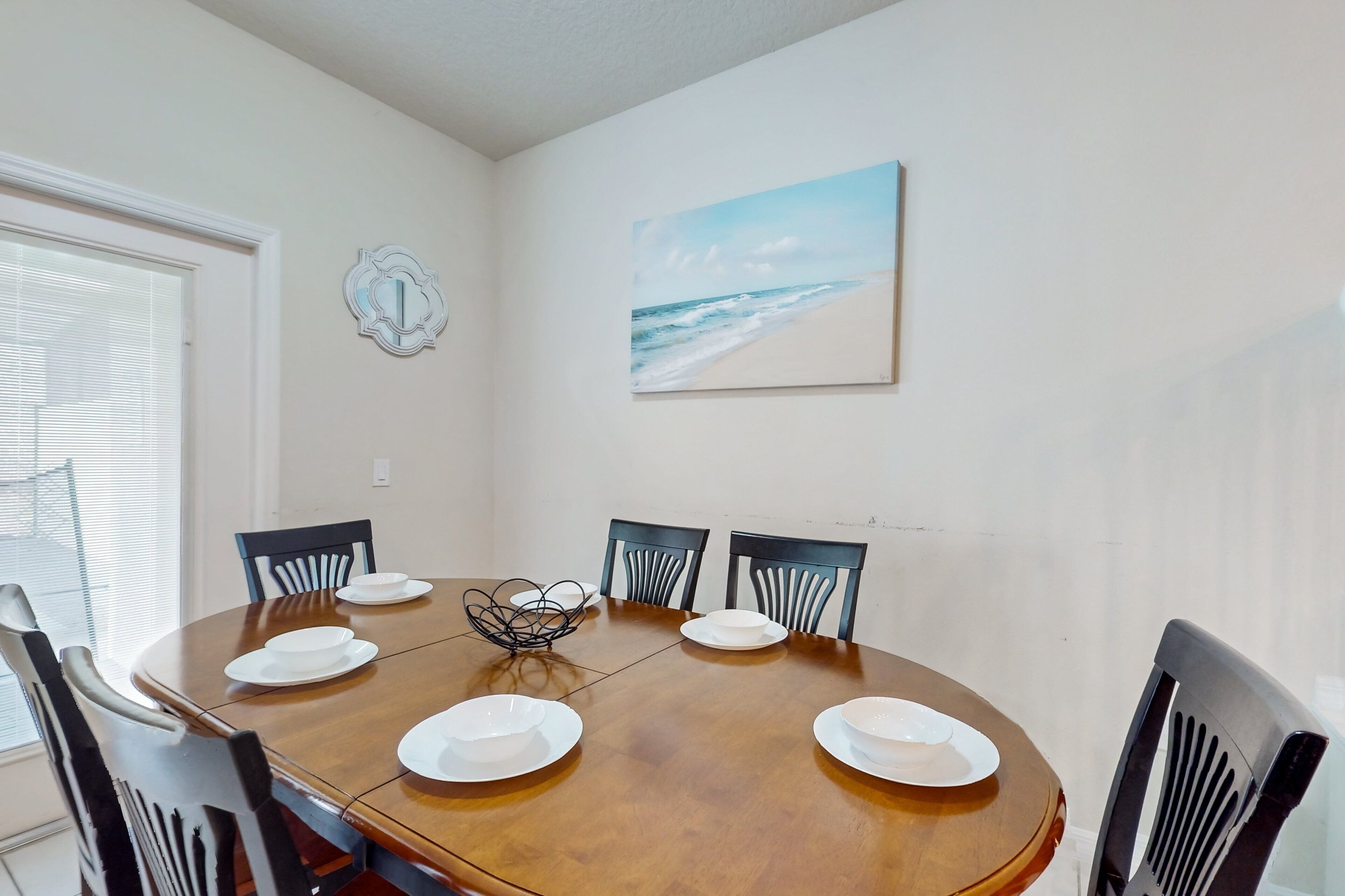Un coin repas avec une table ronde, six chaises et un tableau représentant une plage accroché au mur.