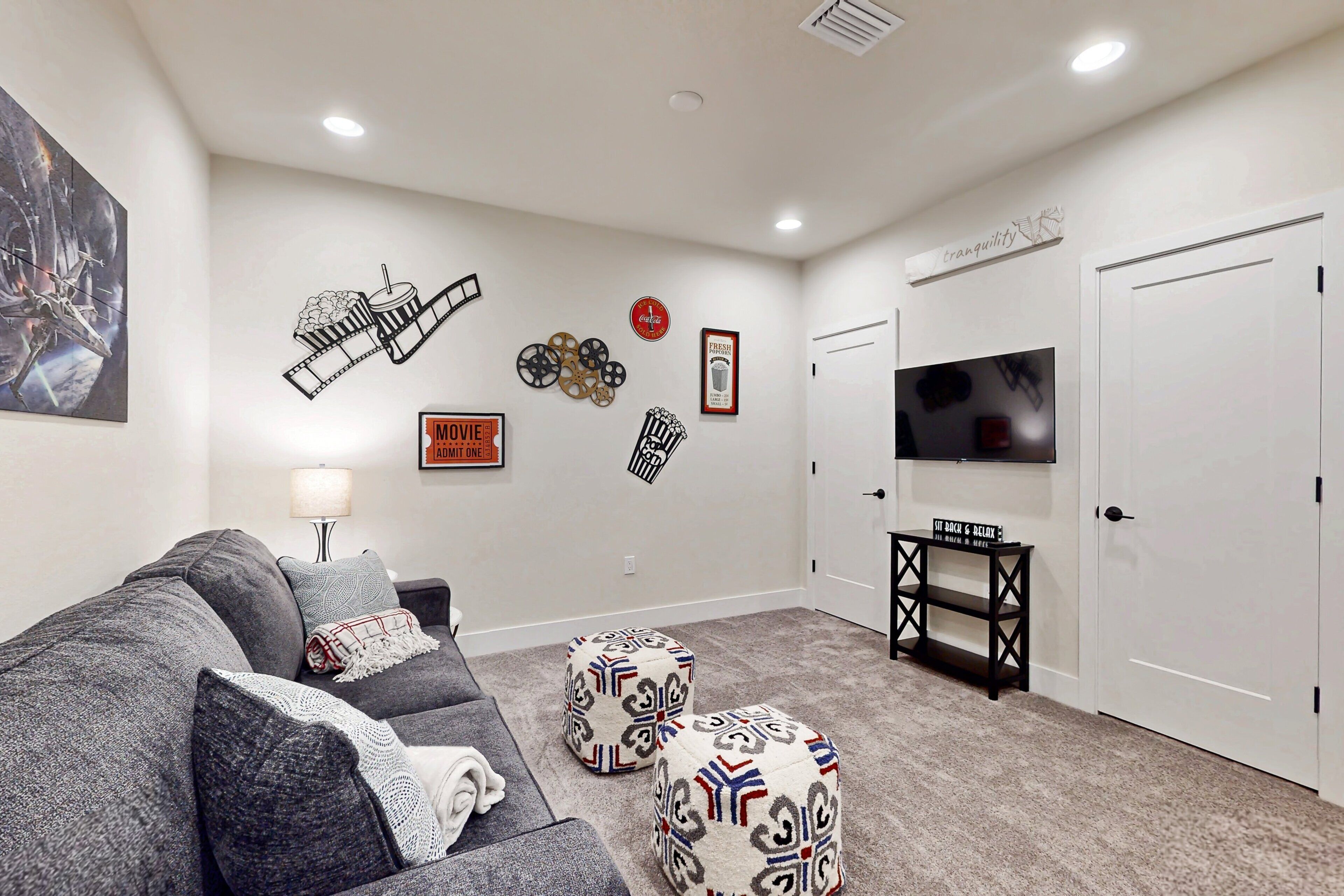 A living room with a grey couch, ottomans, a TV, and wall decorations including a movie sign and a 'FRESH' sign.