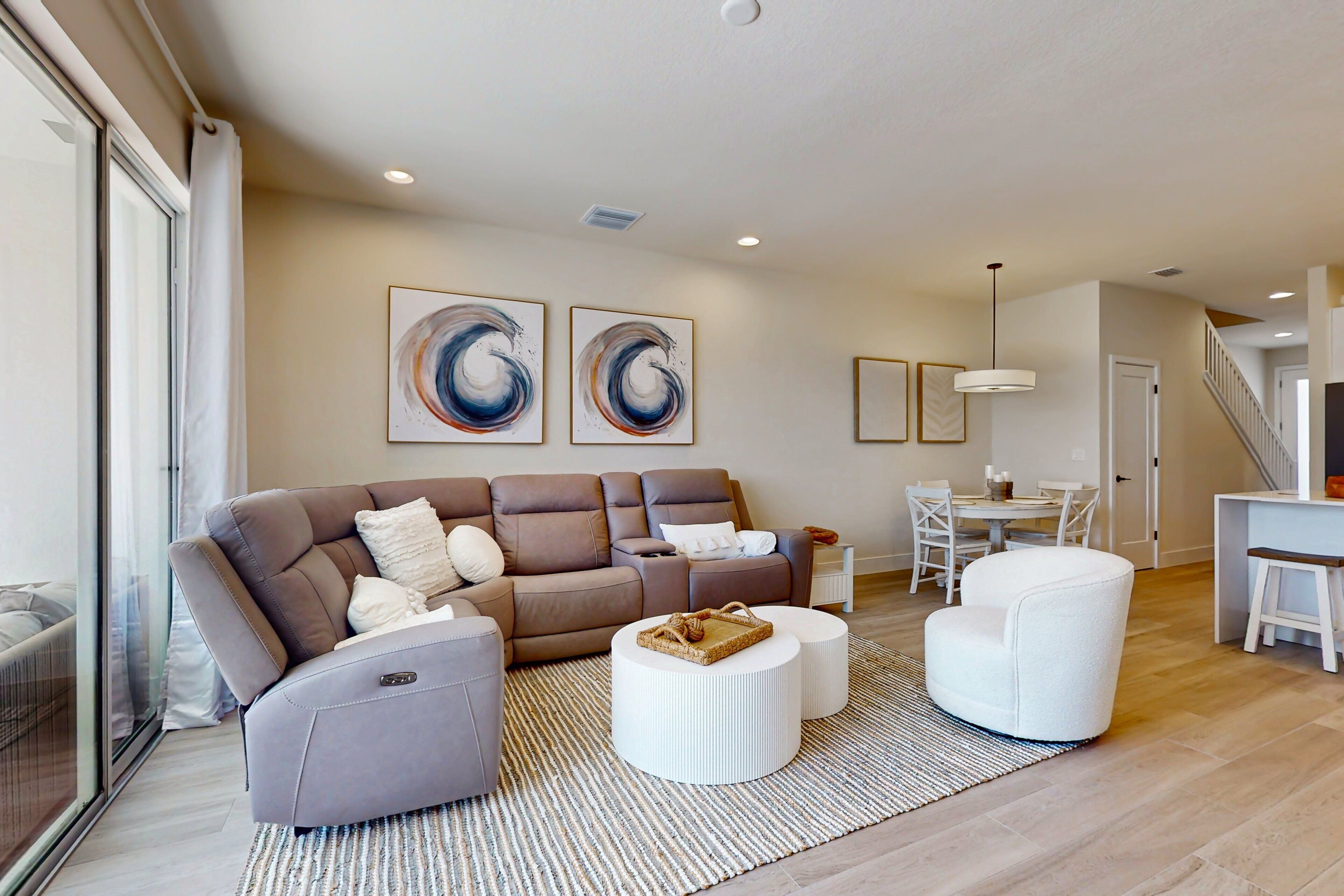 A modern living room with a sectional sofa, a white armchair, and a coffee table. There are abstract paintings on the wall, a dining area with a table and chairs, and a staircase leading to another level.