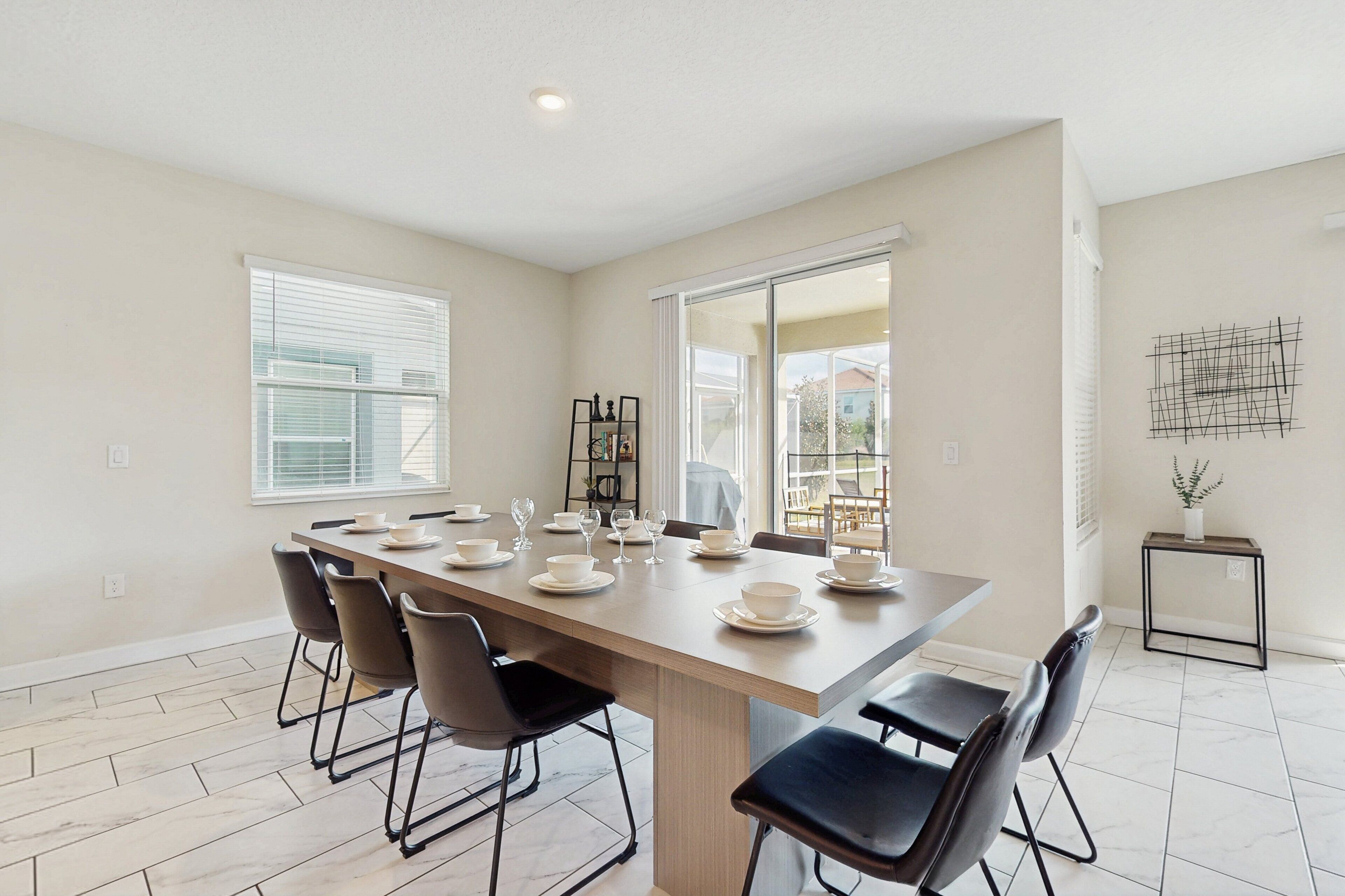 A dining area with a long table set for six, surrounded by chairs, with a view of a house through a glass door.