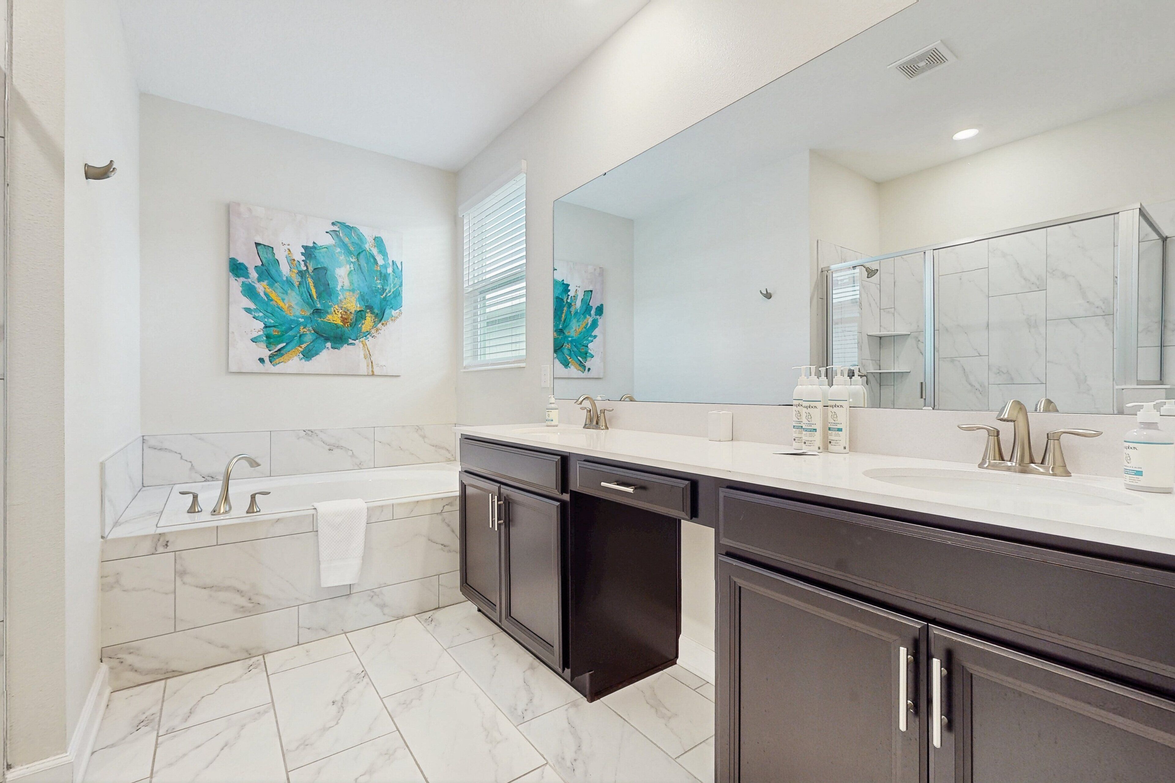 A modern bathroom with a bathtub, two sinks, a large mirror, and artwork on the walls.