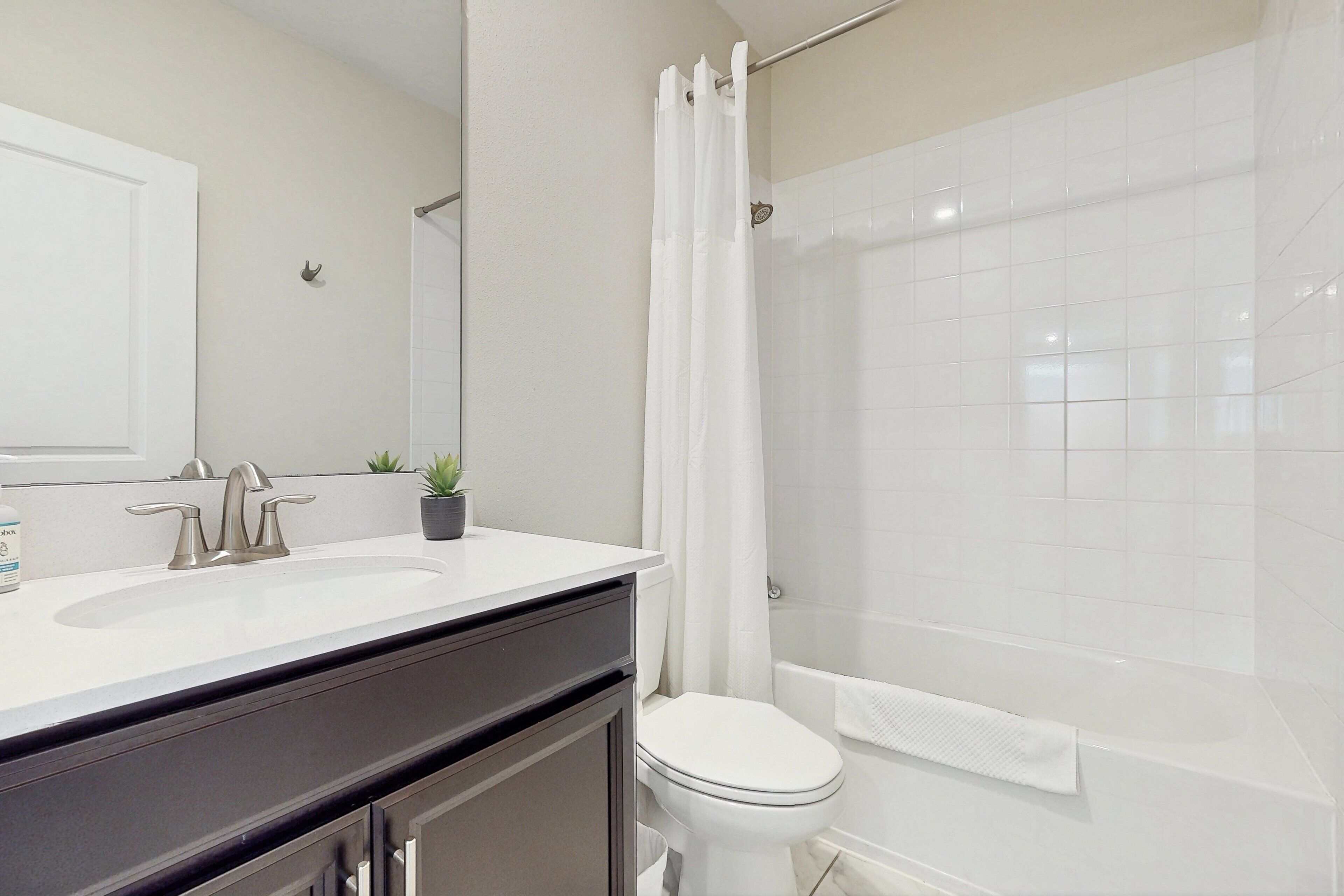 A bathroom with a white sink, a toilet, a shower with a white curtain, and a small potted plant on the sink.