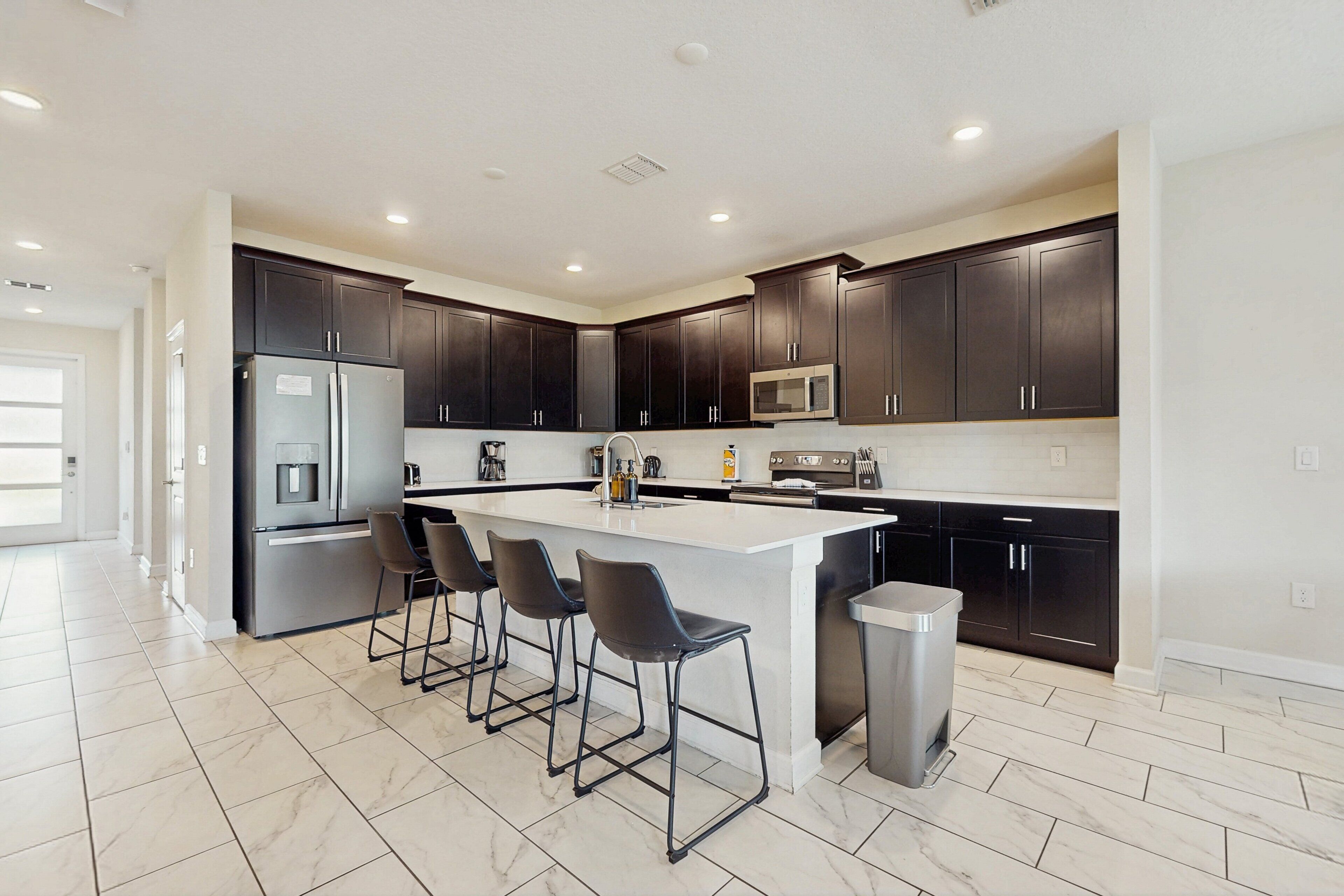 A modern kitchen with dark cabinets, a stainless steel refrigerator, a microwave, and a large island with bar stools.