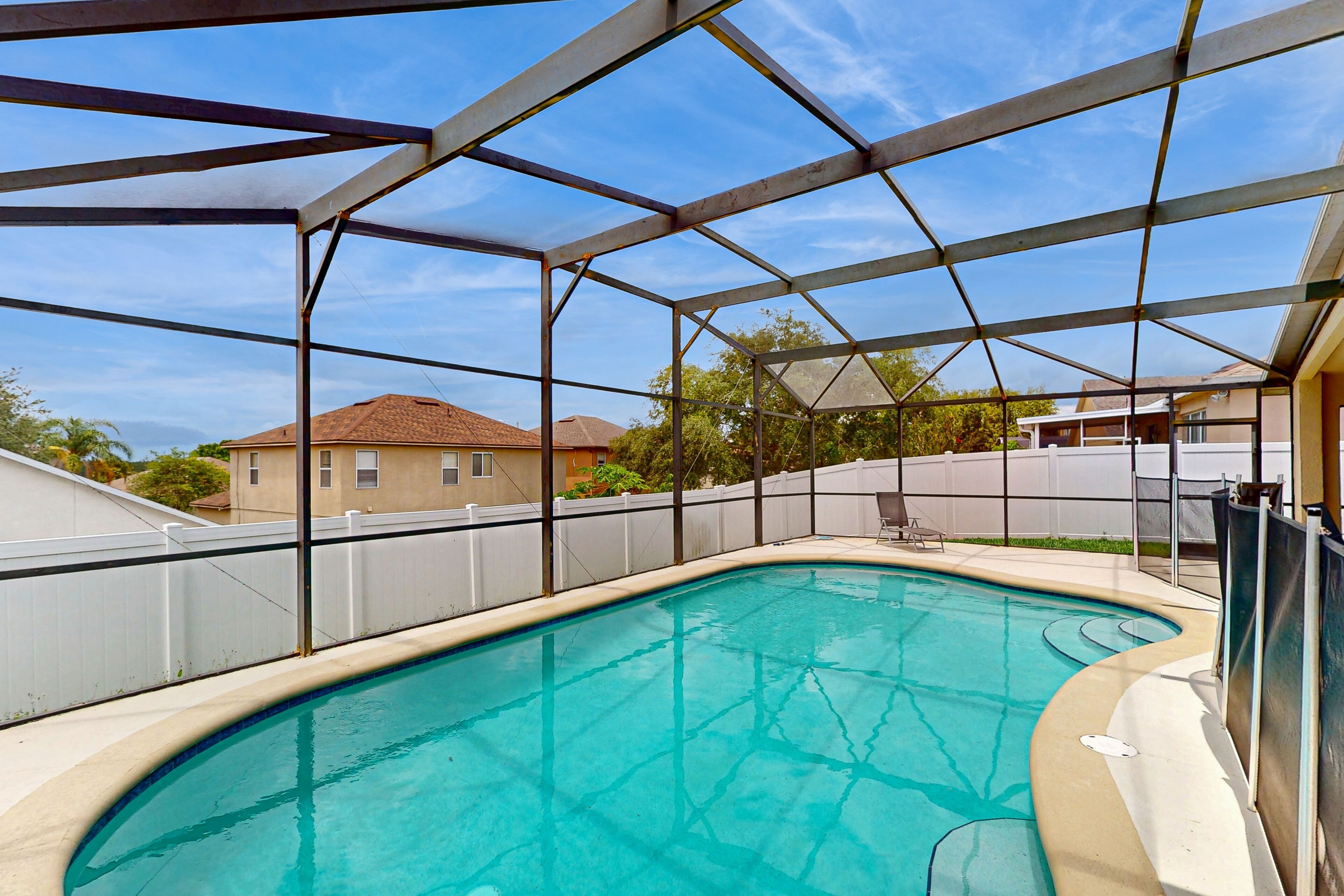 A swimming pool with a metal roof structure, surrounded by a white fence and houses in the background.