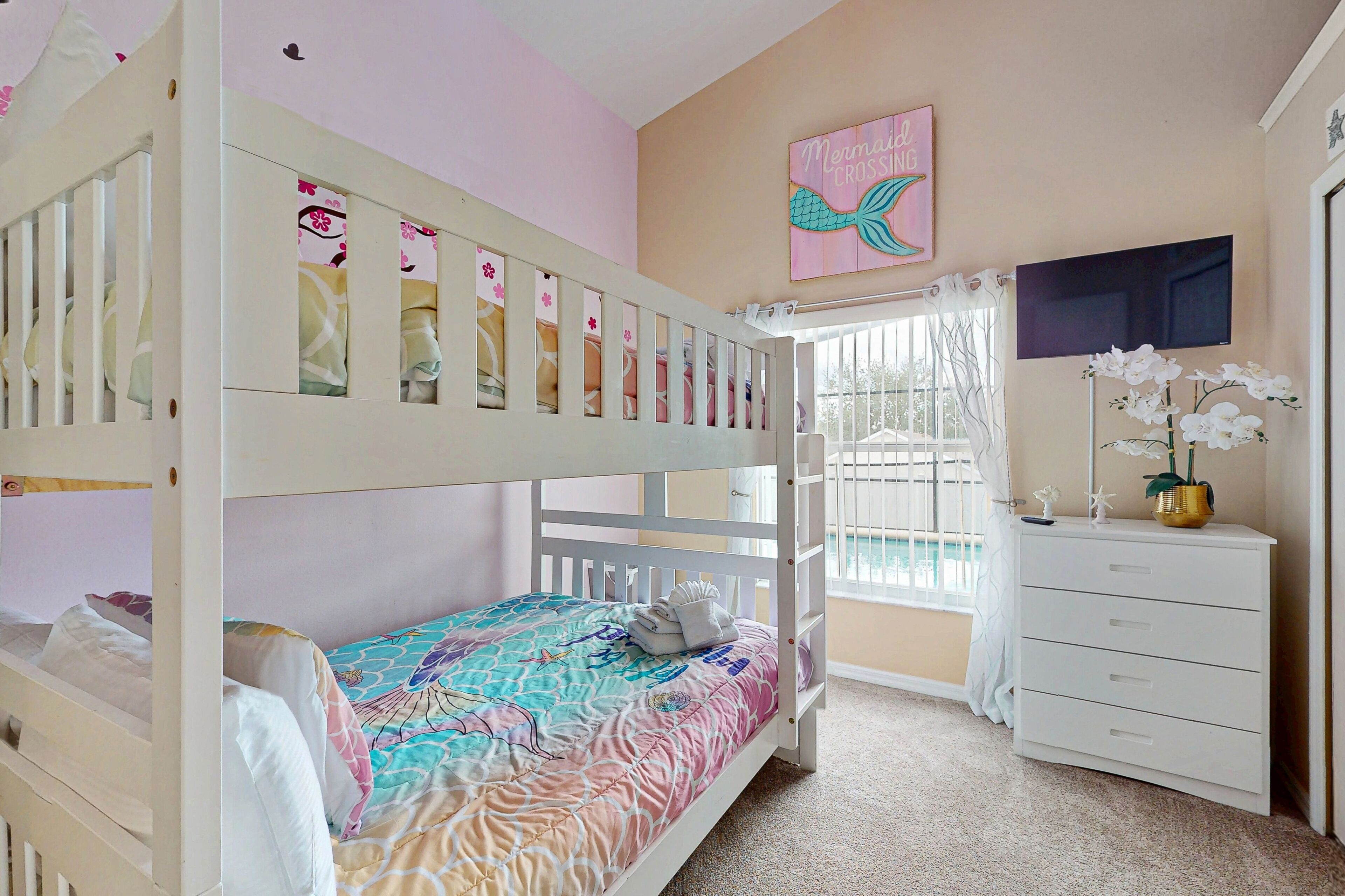 A bunk bed room with a colorful bedspread, a white dresser, a mirror, and a window with curtains.