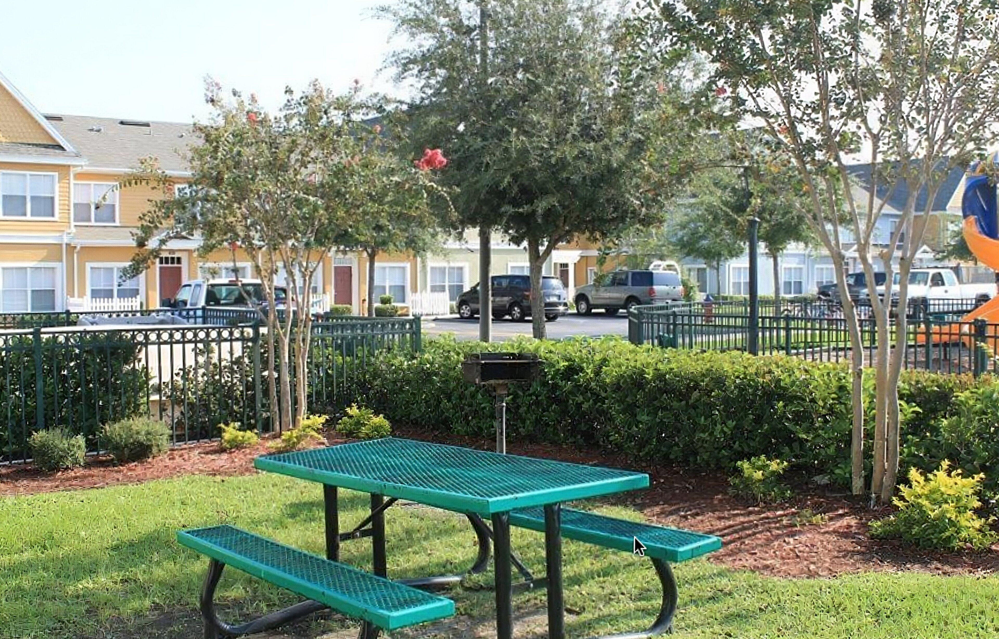 A picnic table with benches, a fenced area, and a row of buildings in the background.