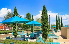 A poolside area with blue umbrellas, green lounge chairs, and tall trees. - Spacious Townhouse With Resort Style Amenities, Waterview and Fitness Center (Kissimmee)