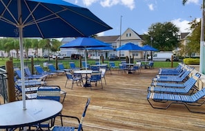 A wooden deck with blue lounge chairs and tables, umbrellas, and a building in the background. - Spacious Townhouse With Resort Style Amenities, Waterview and Fitness Center (Kissimmee)