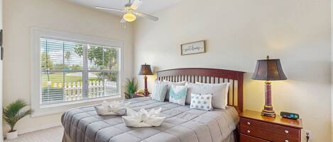 A bedroom with a large bed, two bedside tables with lamps, a ceiling fan, a window with blinds, and a small plant.