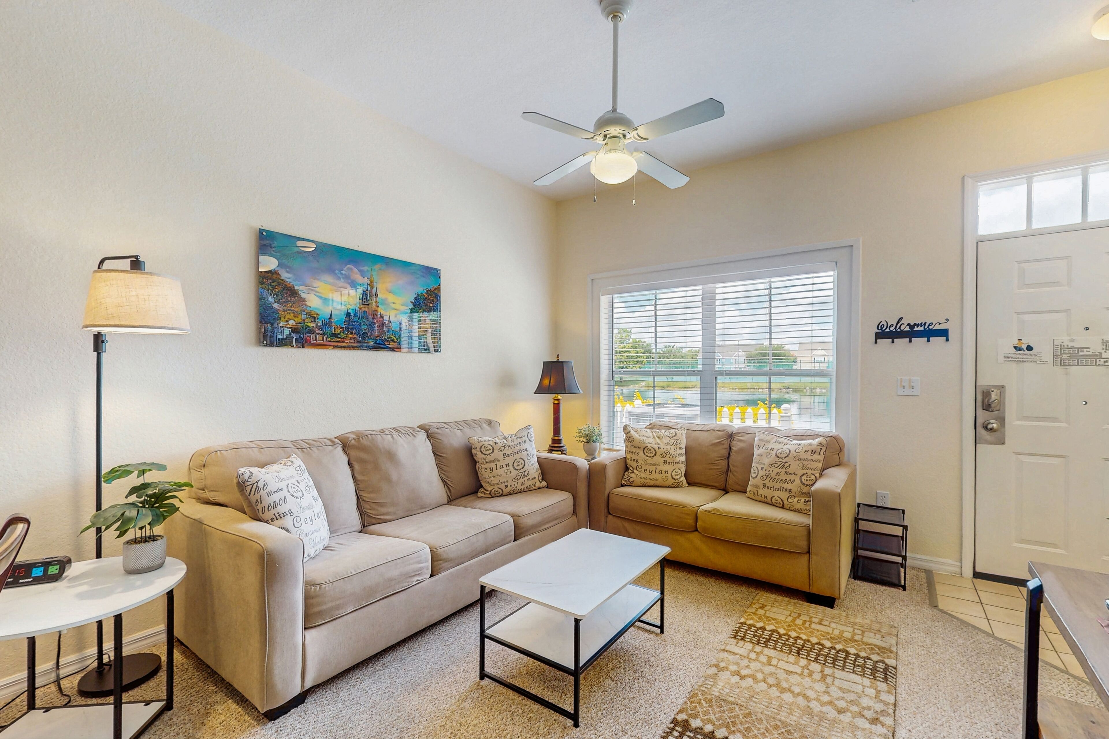 A living room with two beige sofas, a white coffee table, a floor lamp, a painting, and a ceiling fan.