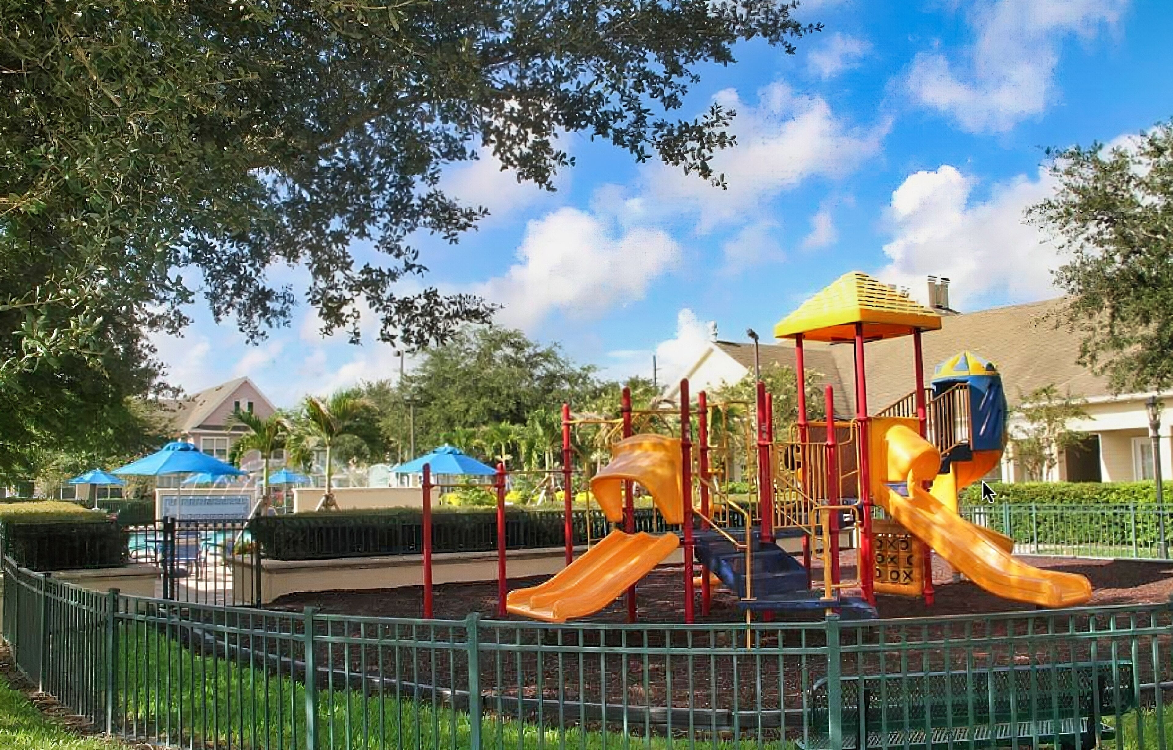 Un parque infantil con toboganes y estructuras para escalar, rodeado por una valla y árboles.