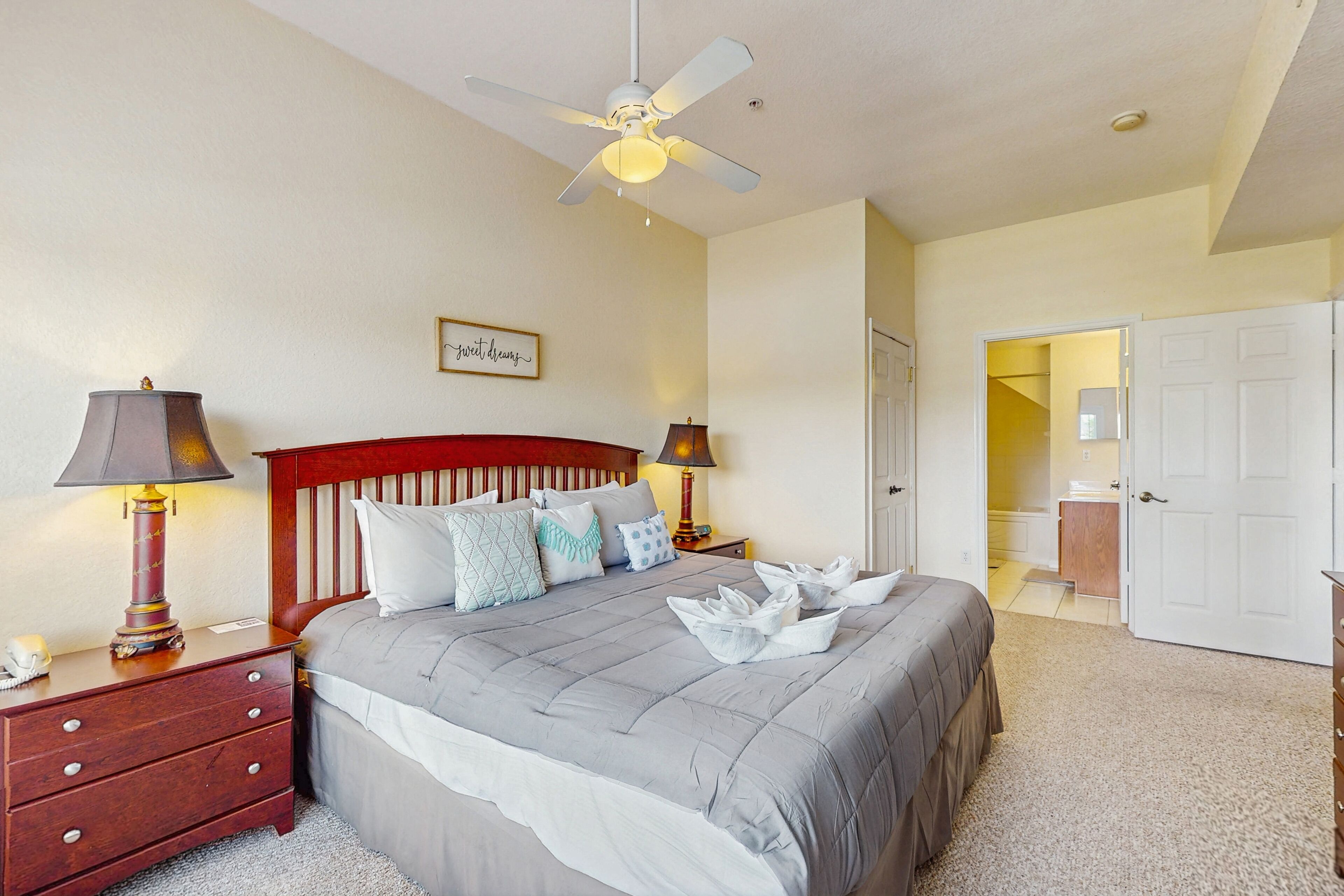 A neatly arranged bedroom with a bed, two bedside tables, a ceiling fan, and a door leading to another room.