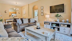 A living room with a sofa, coffee table, TV, and a kitchen area in the background. - Friendly Condo With Pool and hot tub Access, Wifi, Central AC, and Private W/D (Kissimmee)