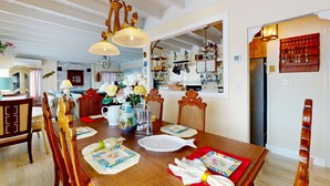 Un comedor con una mesa de madera dispuesta para cuatro personas, rodeada de sillas de madera. Sobre la mesa, hay una araña, y se observa una zona de cocina al fondo.