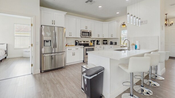 A modern kitchen with white cabinetry, stainless steel appliances, a central island with bar stools, and a visible trash bin. - Water View Retreat With Lanai, Heated Pool, Pool Spa, Game Room, & AC (Kissimmee)