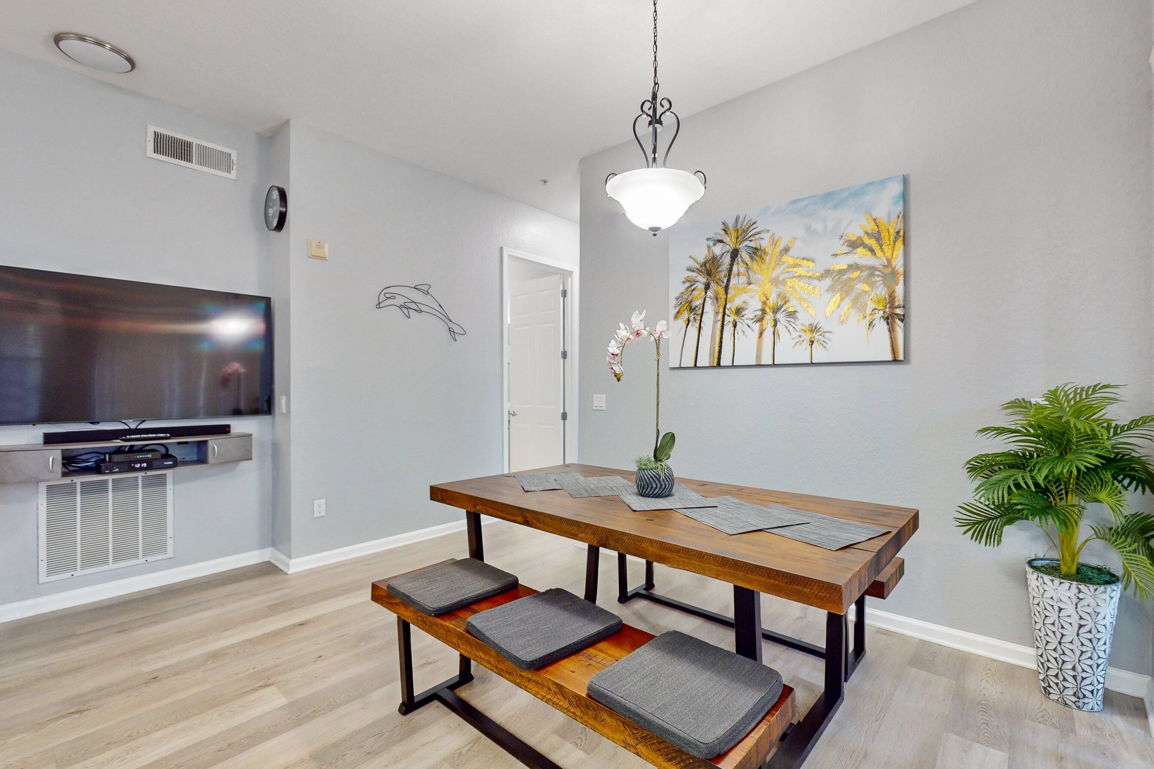 A modern living room with a wooden dining table, bench seating, a flat-screen TV, and a decorative painting.