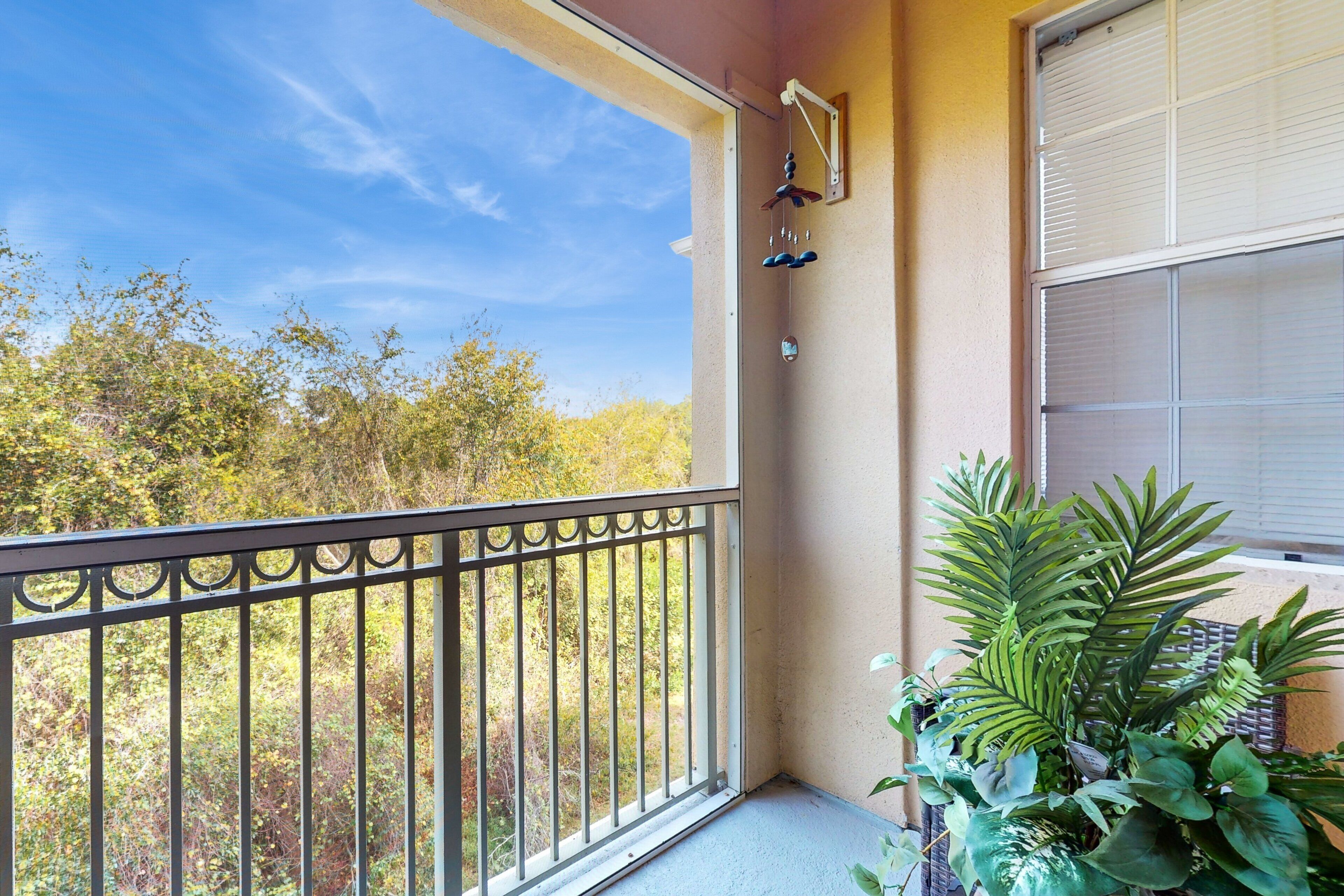 A balcony with a view of trees, a window with blinds, and a hanging wind chime.