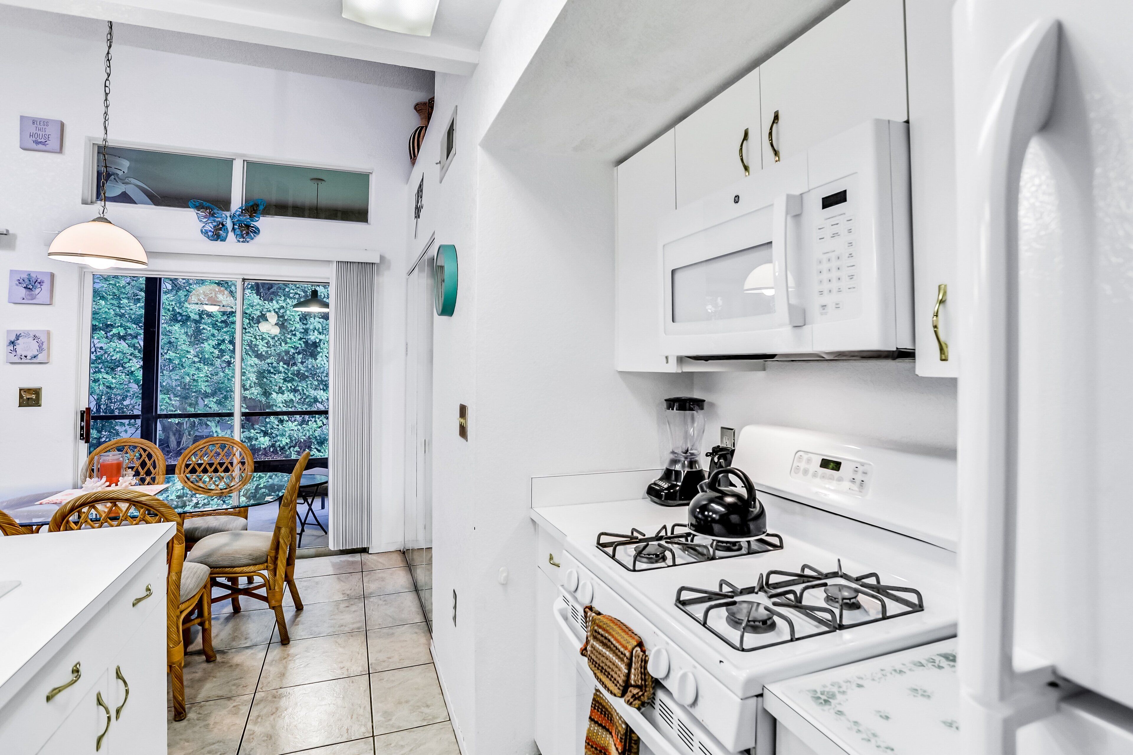 A modern kitchen with white appliances, a dining area with wicker chairs, and a view of greenery outside.