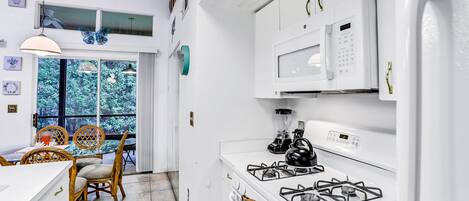 A modern kitchen with white appliances, a dining area with wicker chairs, and a view of greenery outside.