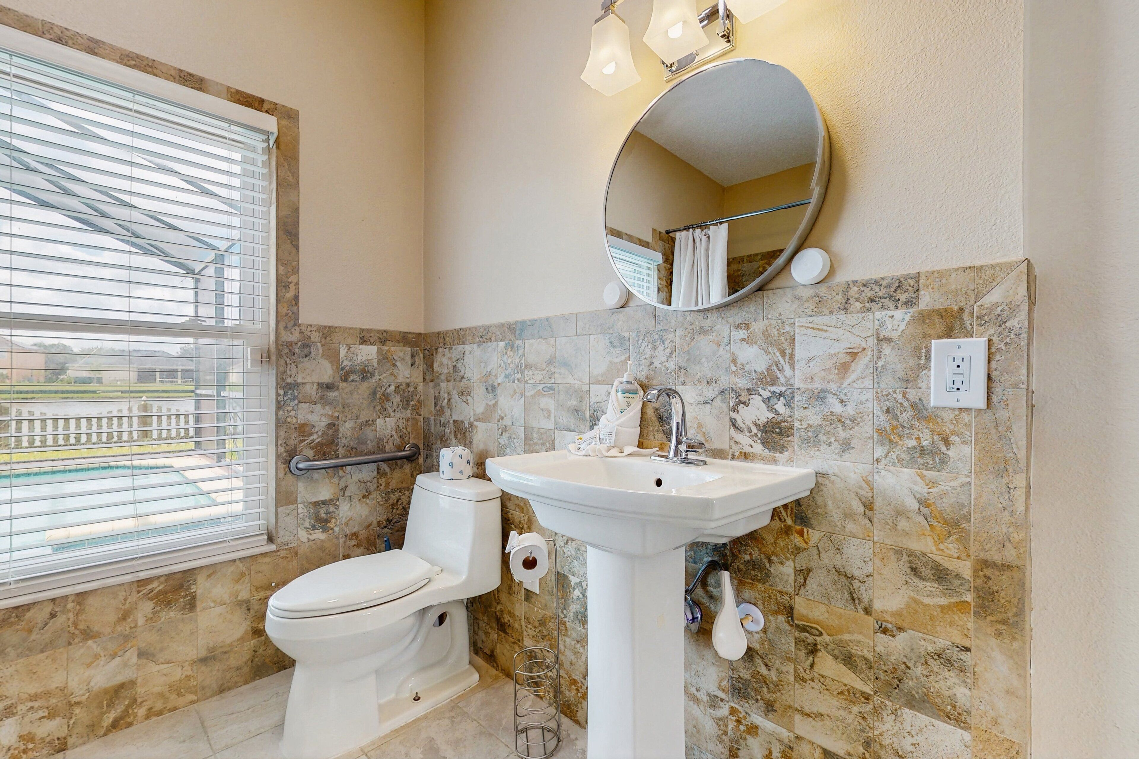 Une salle de bain équipée de toilettes, d’un lavabo, d’un miroir et d’une fenêtre avec des stores.