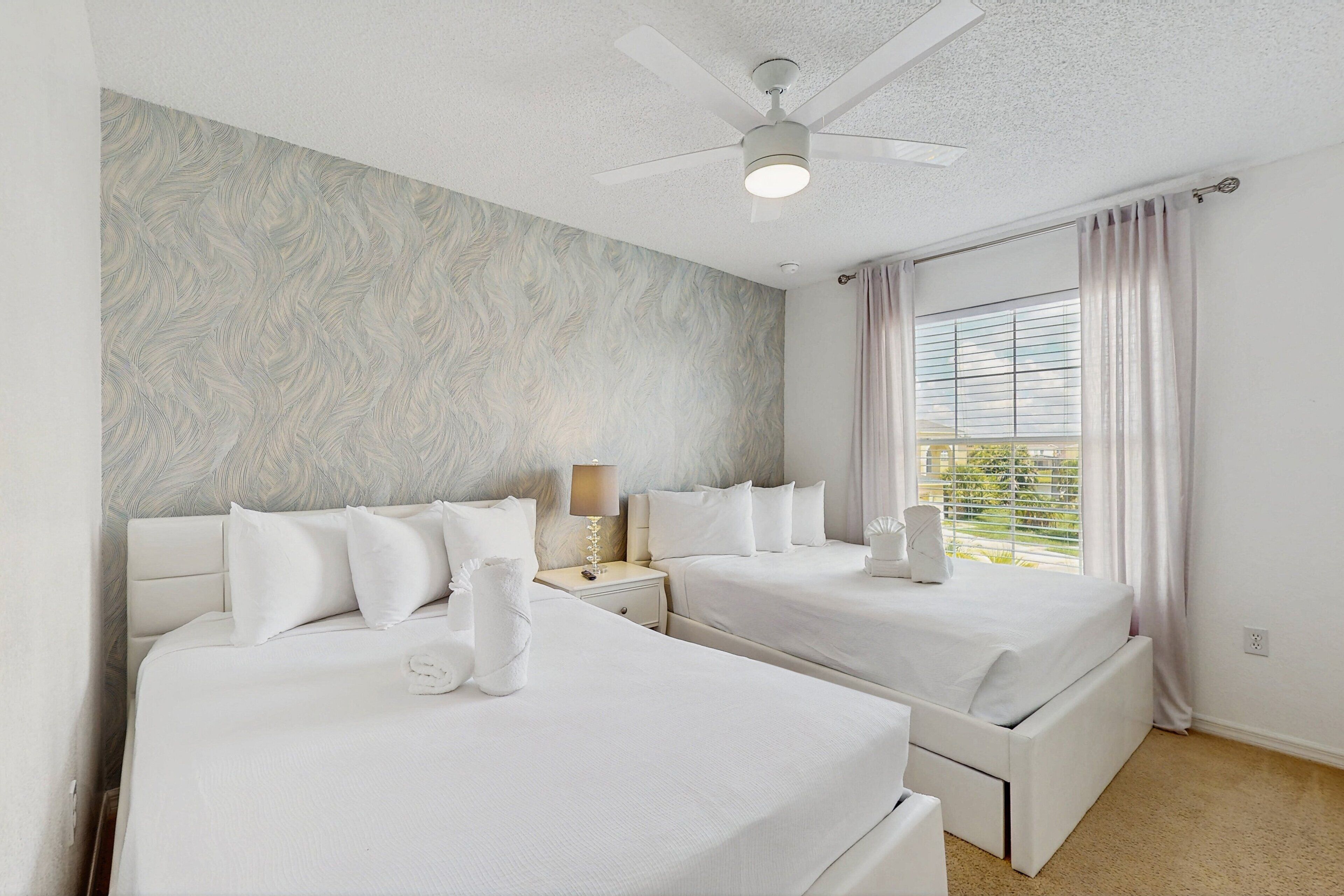 A hotel room with two beds, a ceiling fan, a window with curtains, and a wall with a textured pattern.