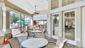 An outdoor patio area with round tables and chairs, a ceiling fan, and large windows offering views of the pool and outdoor seating. - Resort Condo Near Disney With Pool, hot Tub, Tennis, Central AC, & Washer/dryer (Kissimmee)