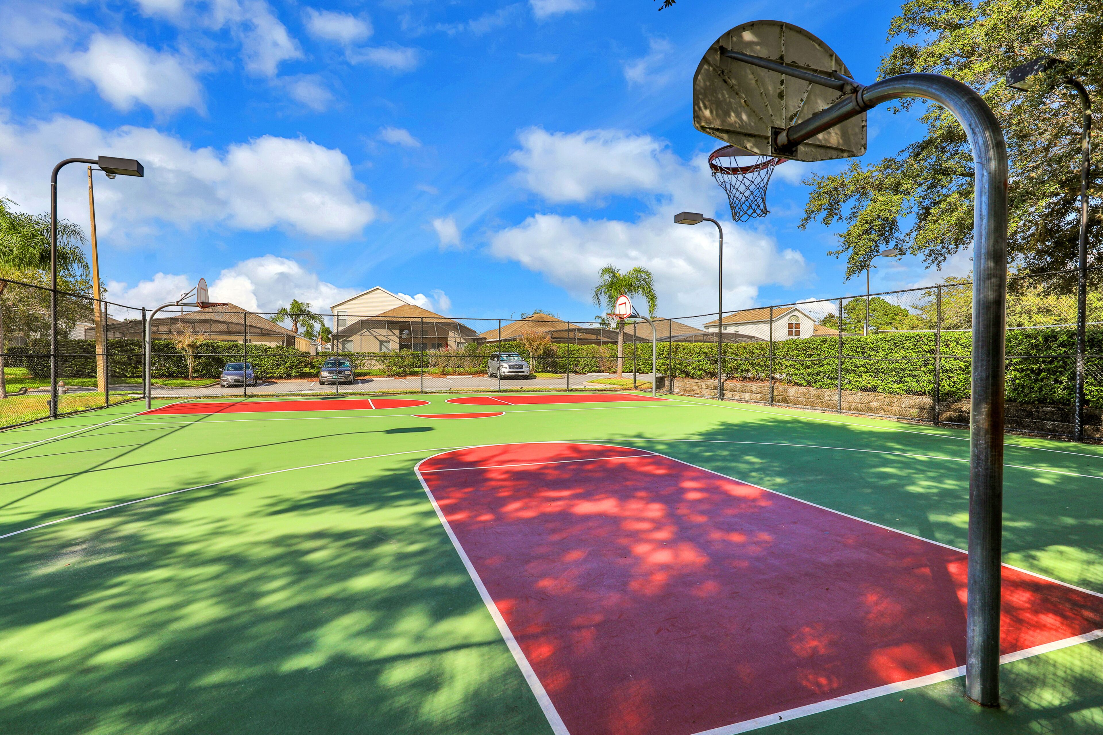 En udendørs basketballbane med grøn og rød spilleflade, omgivet af et hegn, under en klar himmel.
