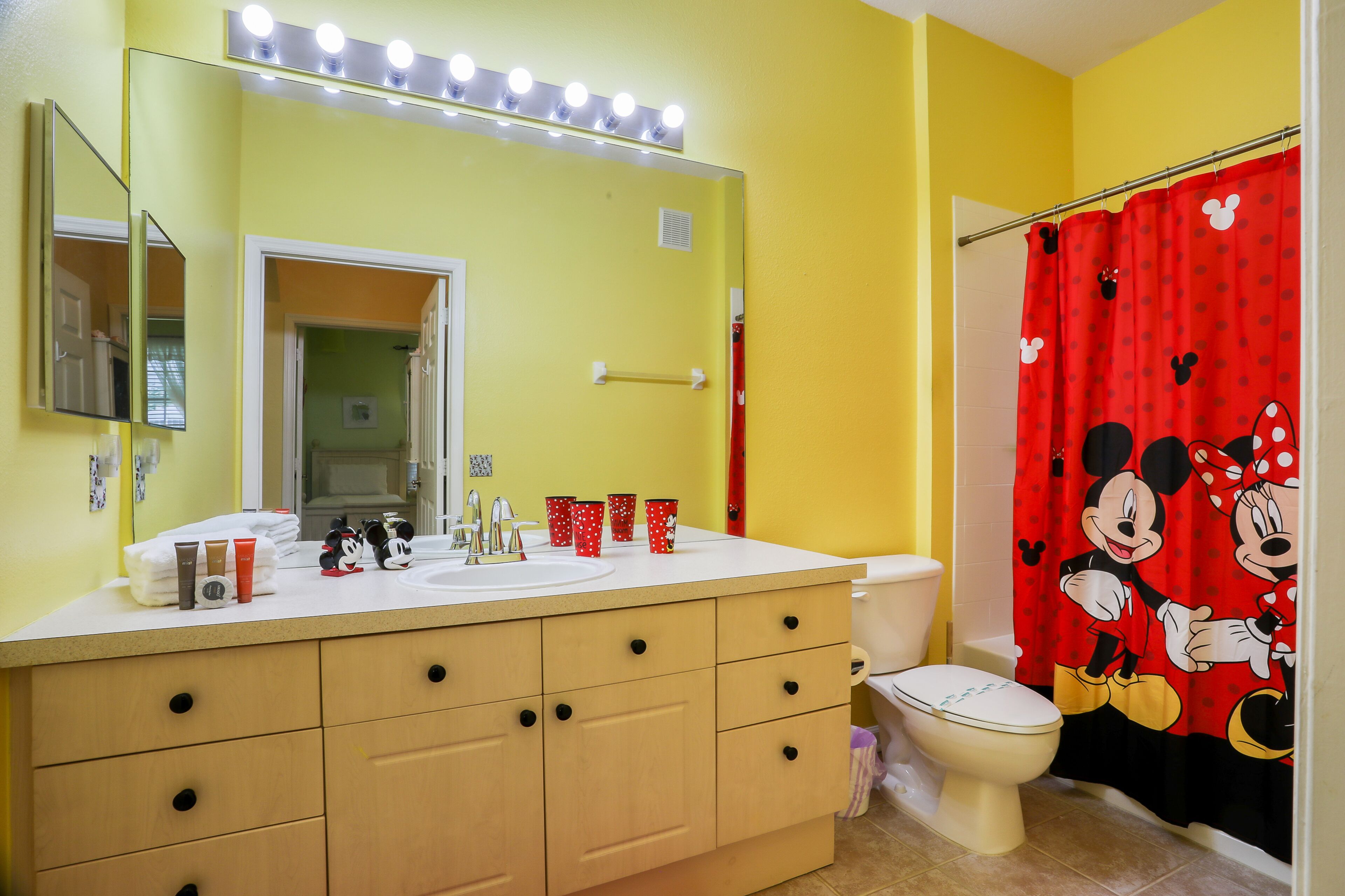 A bathroom with a yellow wall, a white sink, a mirror with lights, a Mickey Mouse shower curtain, and a toilet.
