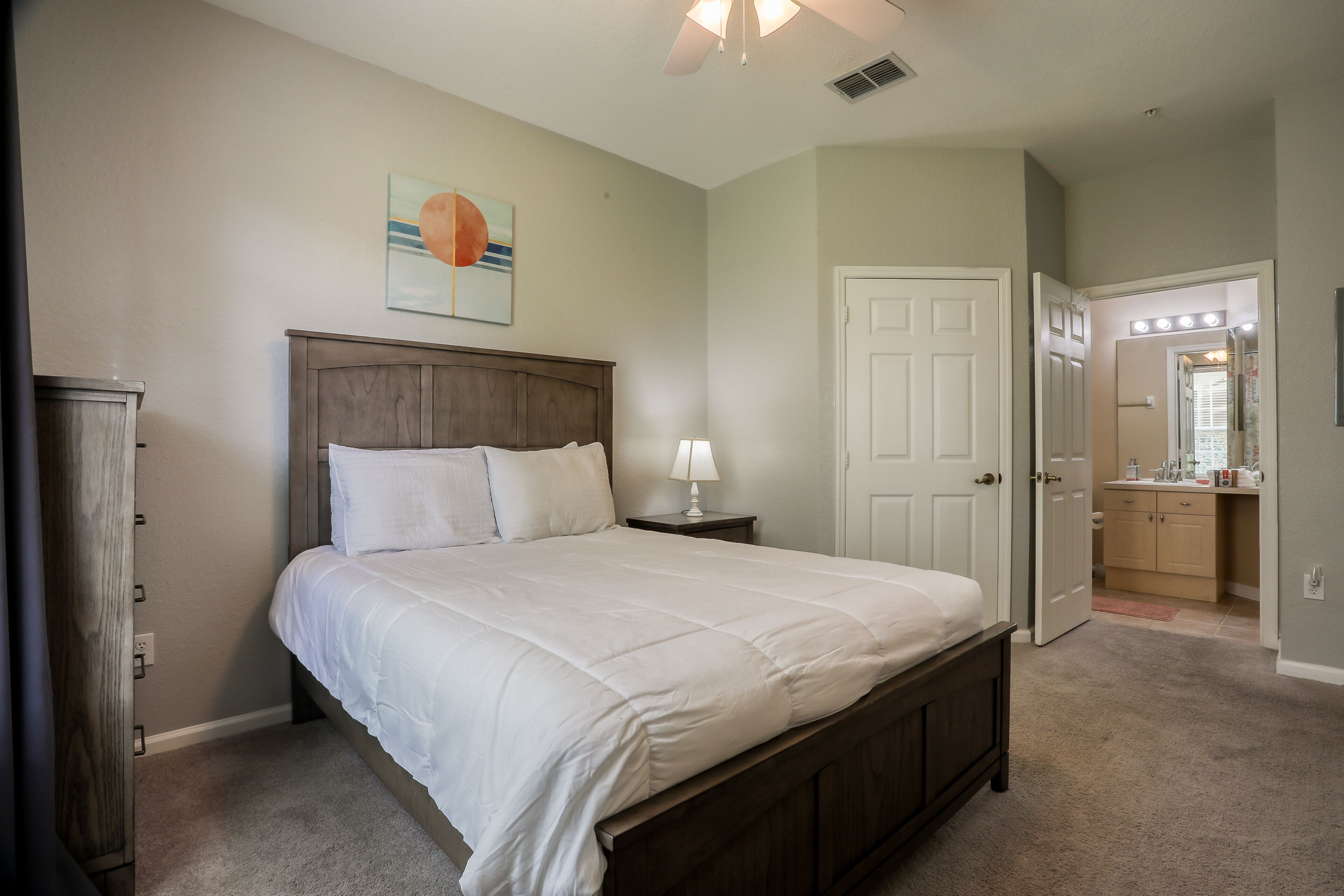 A bedroom with a bed, bedside table, and a nightstand. There is a door leading to a bathroom with a mirror and lights.