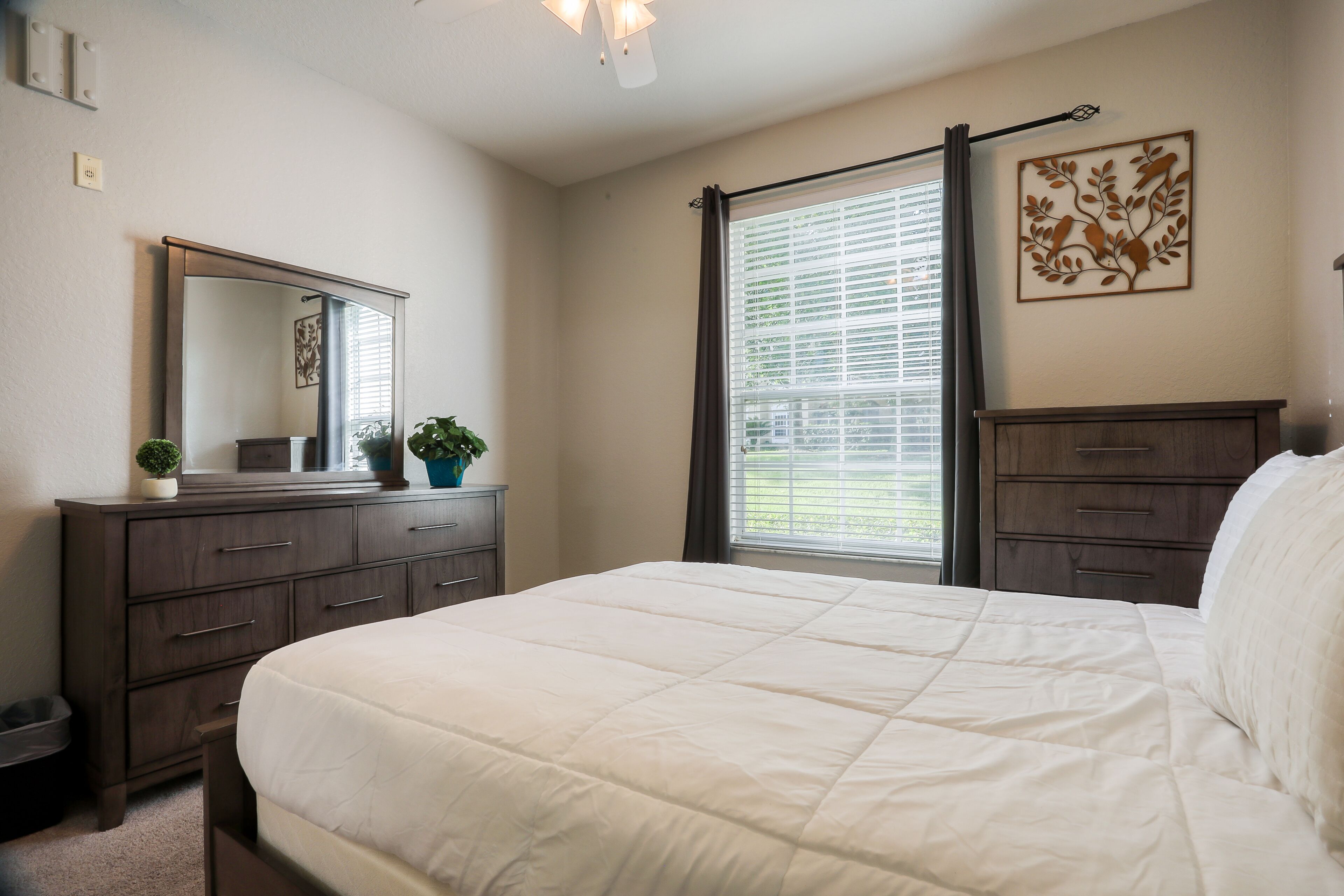A bedroom with a large bed, a dresser, a mirror, and a window with blinds.