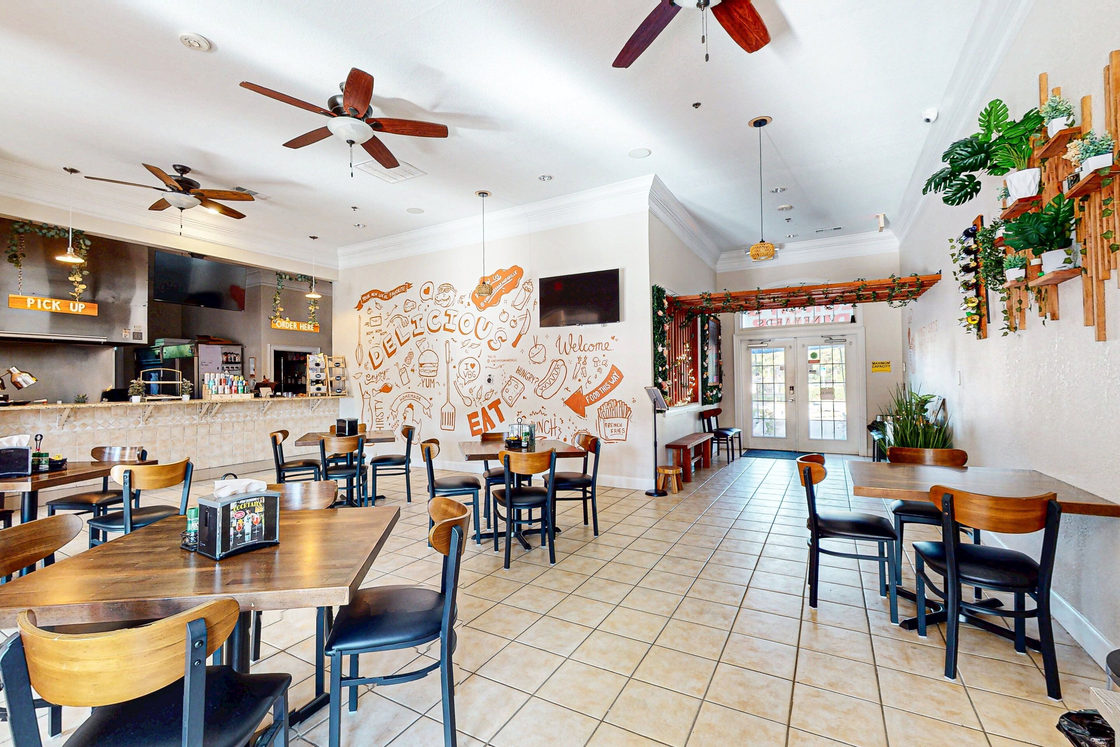 Un restaurant doté de nombreuses tables et chaises, de ventilateurs de plafond et d’un mur orné de dessins sur le thème de la nourriture.