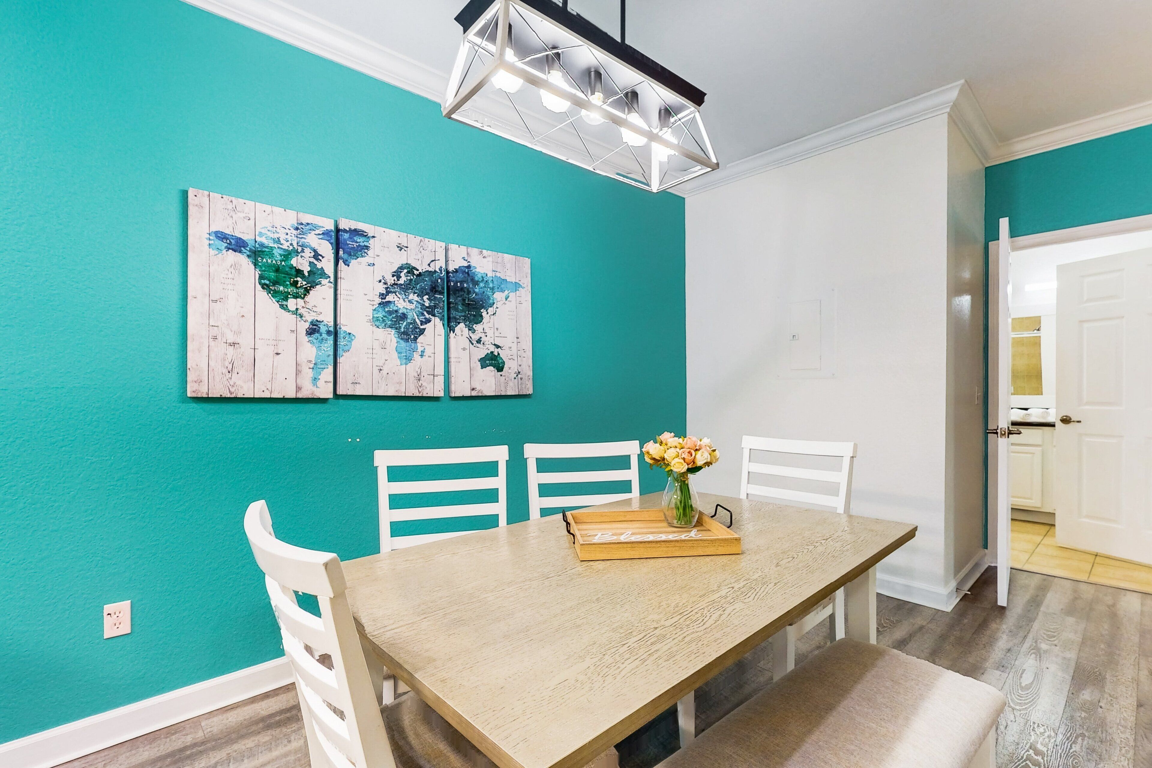 Un comedor con mesa de madera, sillas blancas, un cuadro de un mapa mundial y un florero con flores.