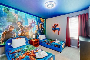 Une chambre avec deux lits, une fresque représentant des super-héros, une commode et une fenêtre avec des rideaux.