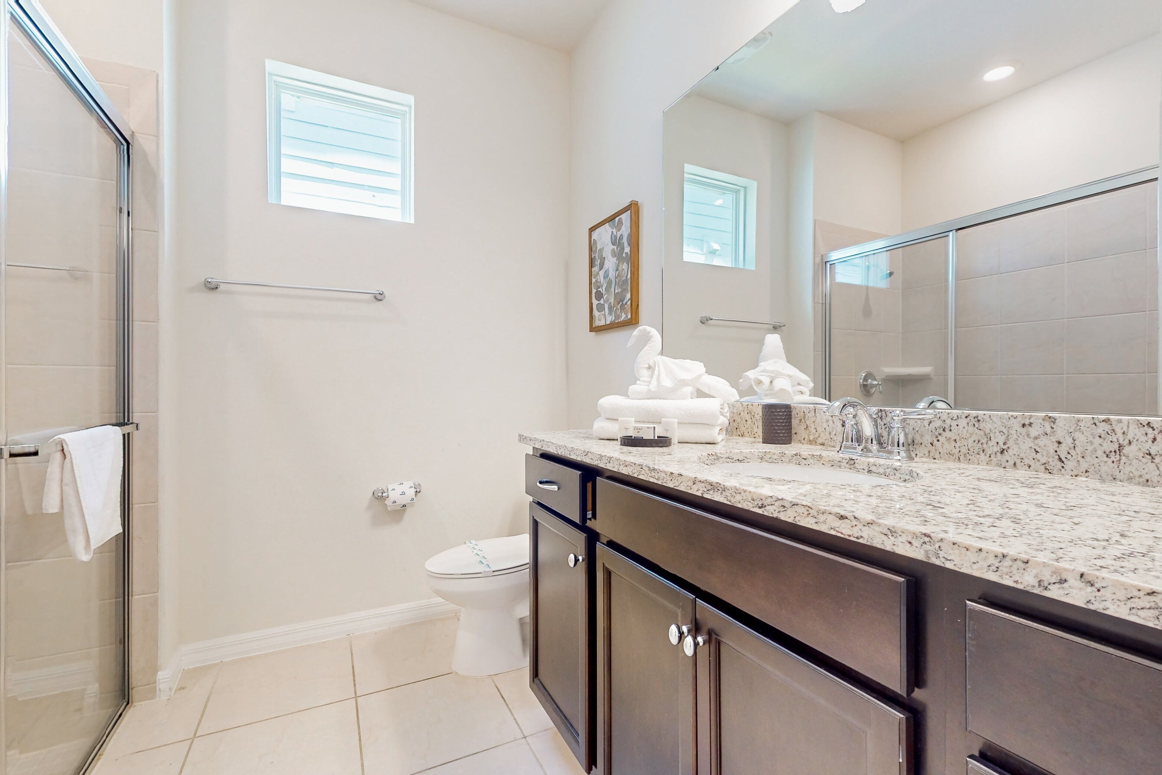 A bathroom with a glass shower enclosure, a white toilet, a large mirror, and a granite countertop.