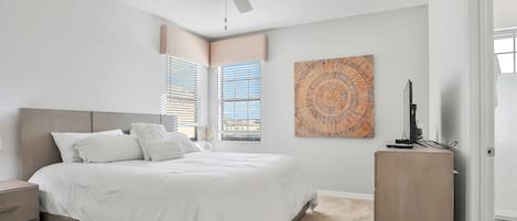 Un dormitorio con cama, ventilador de techo, ventana con persianas, una obra de arte en la pared y un mueble pequeño.