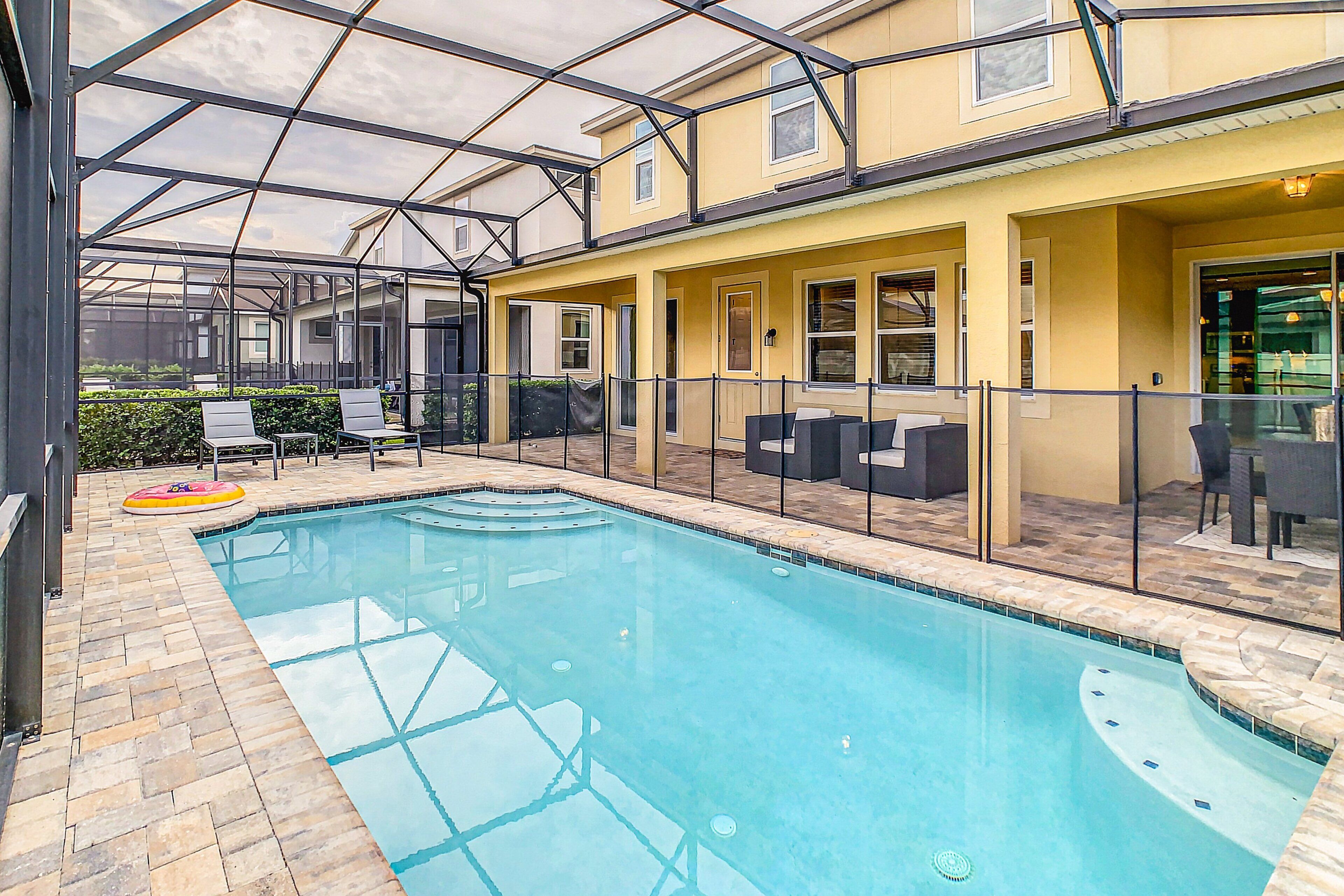 A swimming pool with a glass enclosure, outdoor seating, and a yellow building with multiple windows.