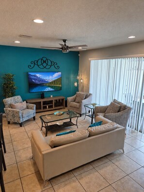 A living room with a teal accent wall, a flat-screen TV, a ceiling fan, and a large window with blinds. - Limited-time Special Discounts Spacious Home w/ Pool and Resort Amenities (Kissimmee)