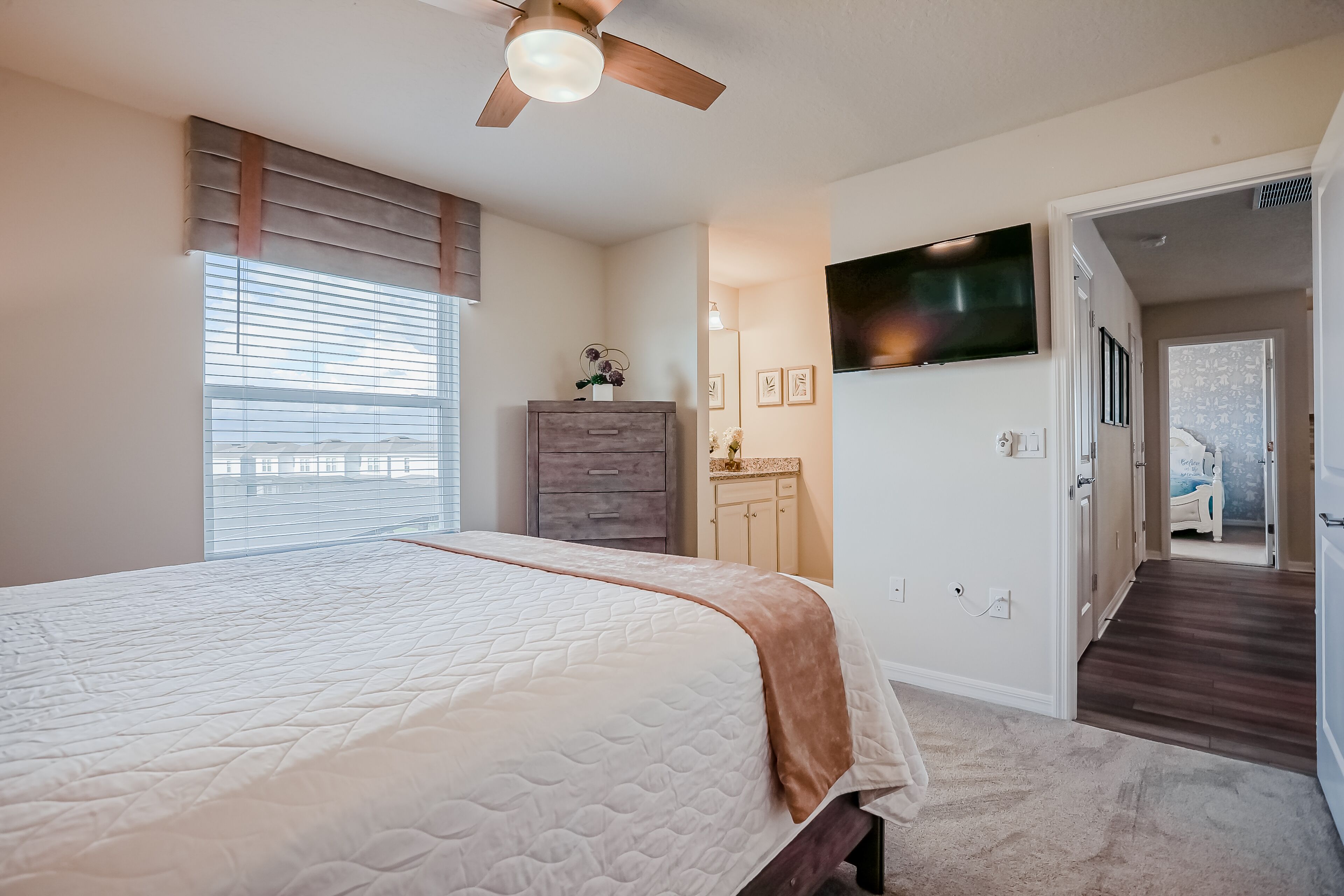 A bedroom with a bed, ceiling fan, window blinds, dresser, vanity, and a TV on the wall.