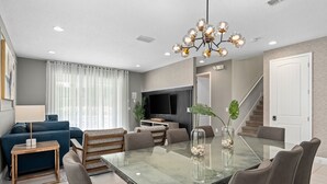 A modern living room with a glass-top dining table, comfortable seating, a TV, and a staircase. - Dog-friendly Getaway With Private Pool + Spa, Putting Green, Patio, & Game Room (Kissimmee)
