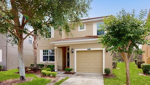 A two-story residential building with a beige facade, a garage, and a well-maintained lawn with small trees and shrubs. - Dog-friendly Home With Private Pool, High-speed Wifi, Central AC, & Washer/dryer (Kissimmee)