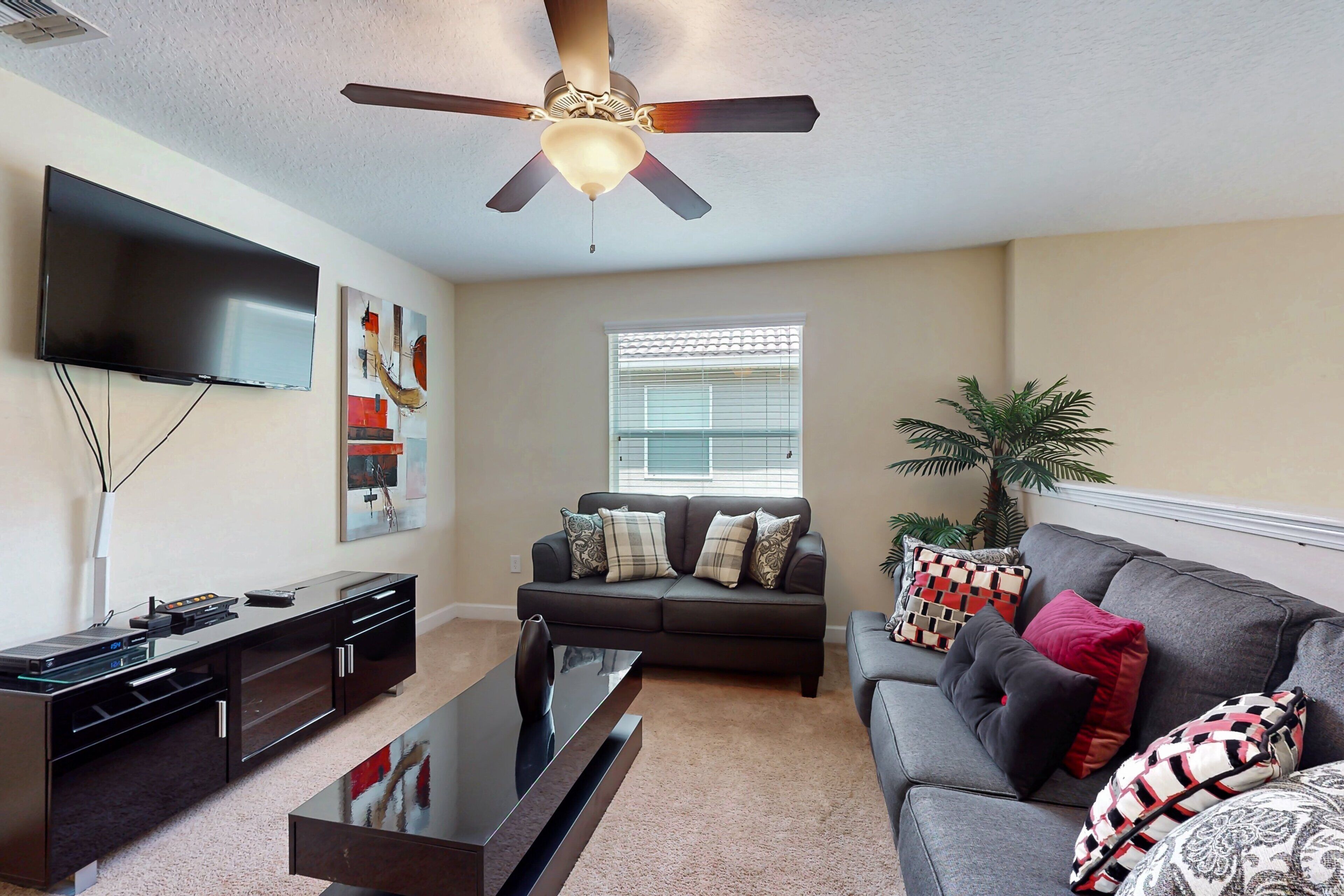 A living room with a flat-screen TV, a ceiling fan, a sofa, a coffee table, and a window with blinds.