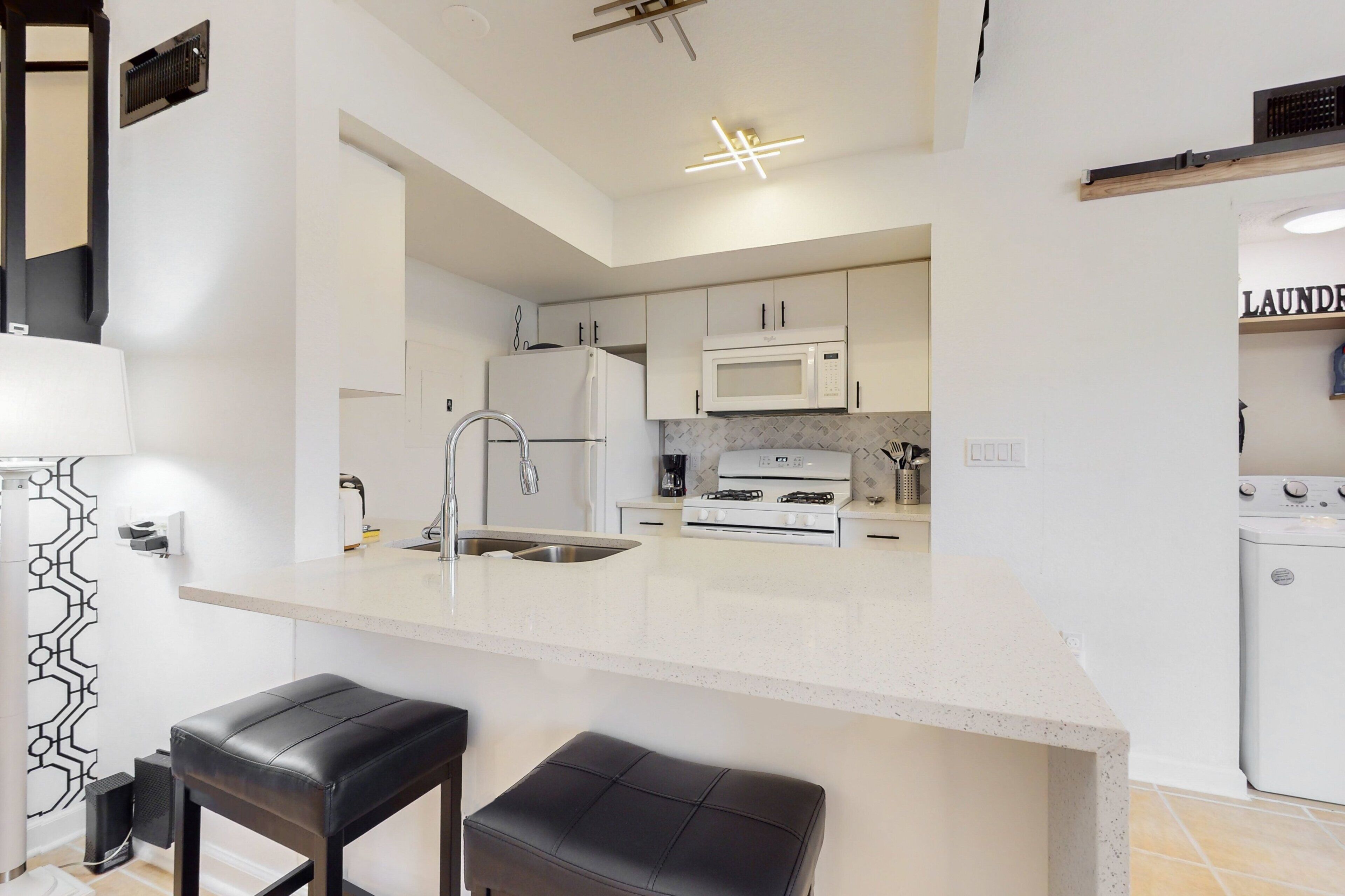 A modern kitchen with white cabinets, a built-in microwave, a gas stove, and a sink. There are two black bar stools at the island.