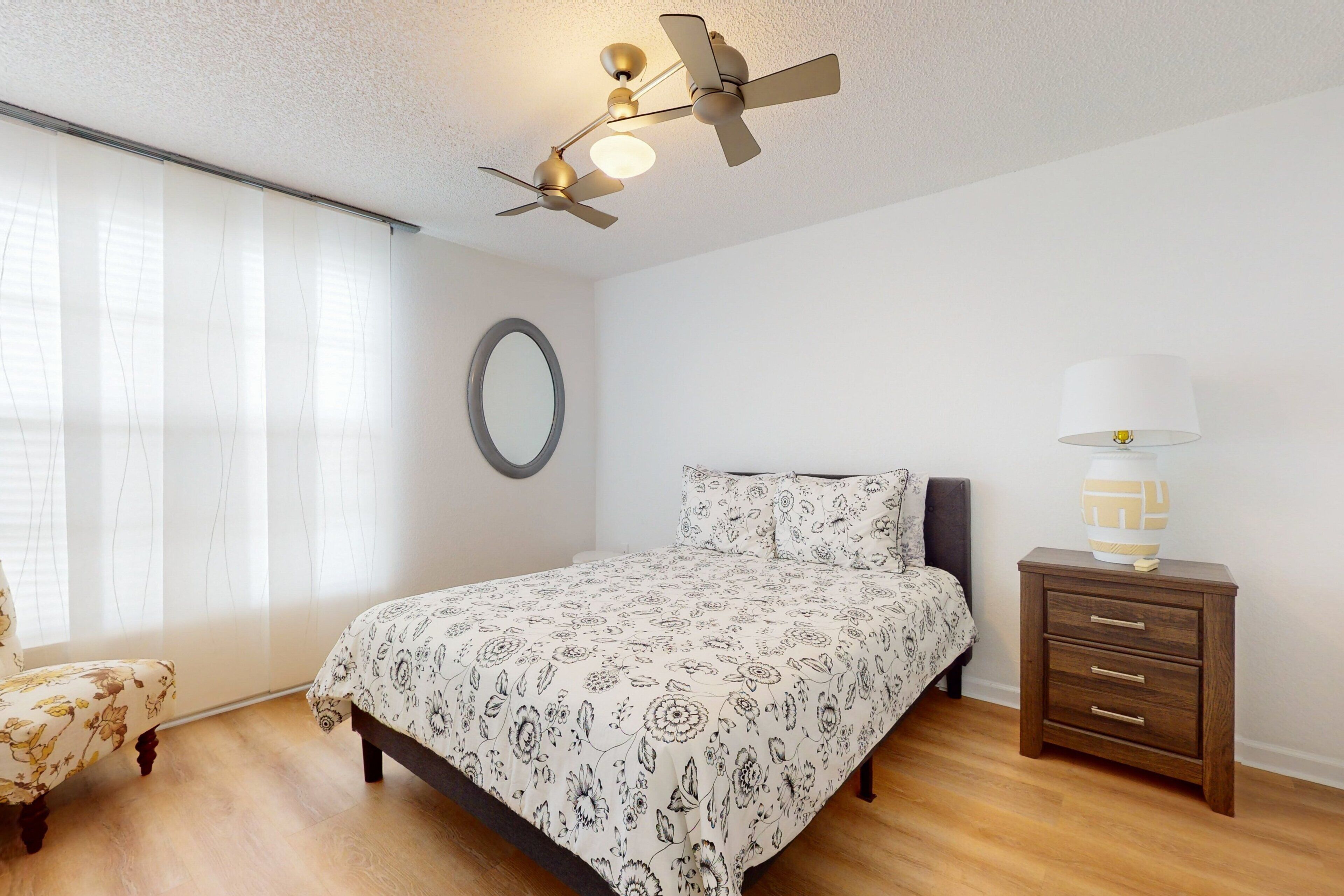 A bedroom with a bed, a bedside table, a lamp, a ceiling fan, and a window with curtains.