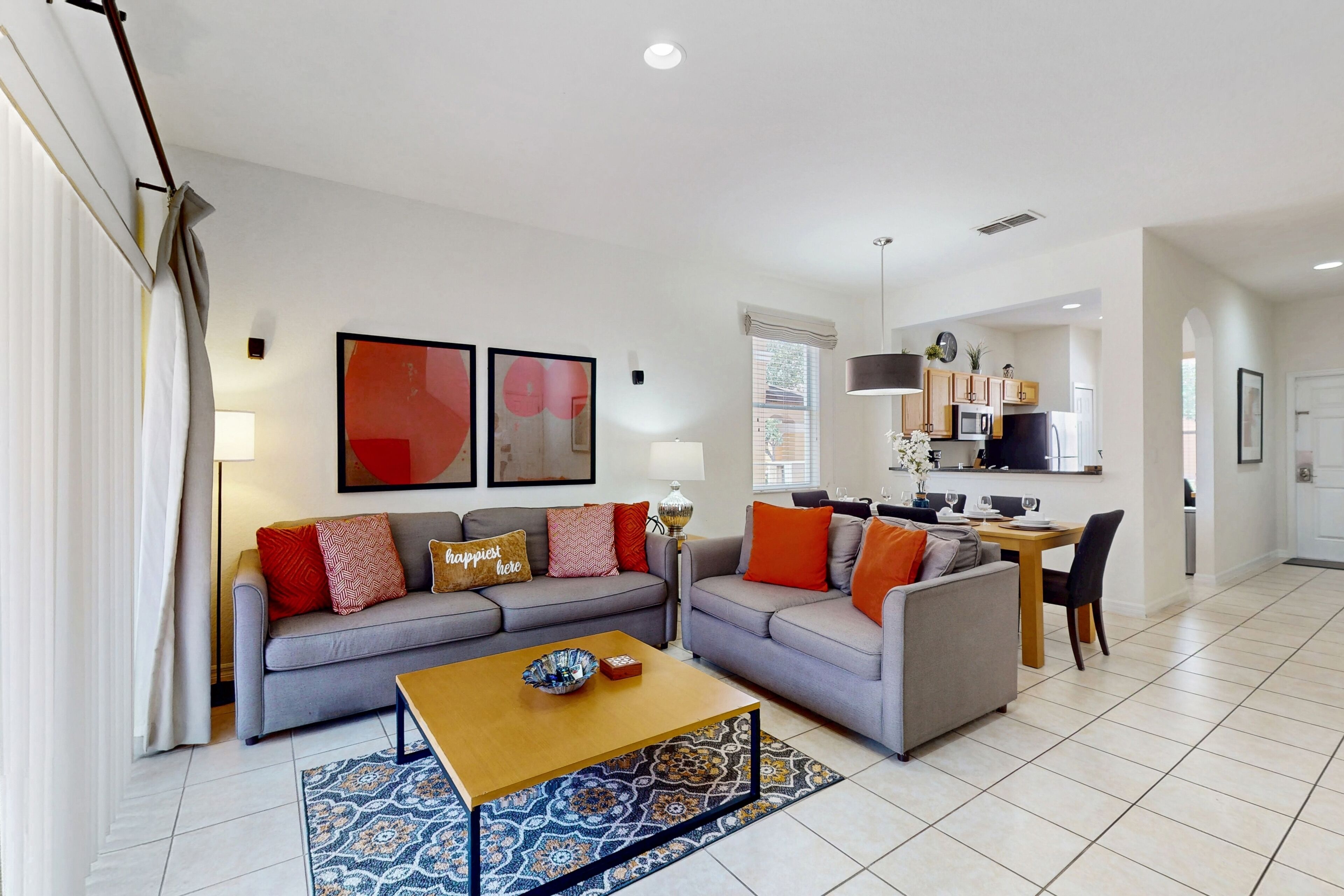 A modern living room with a grey sofa, a wooden coffee table, and a patterned rug. A dining area with a table and chairs is visible in the background.