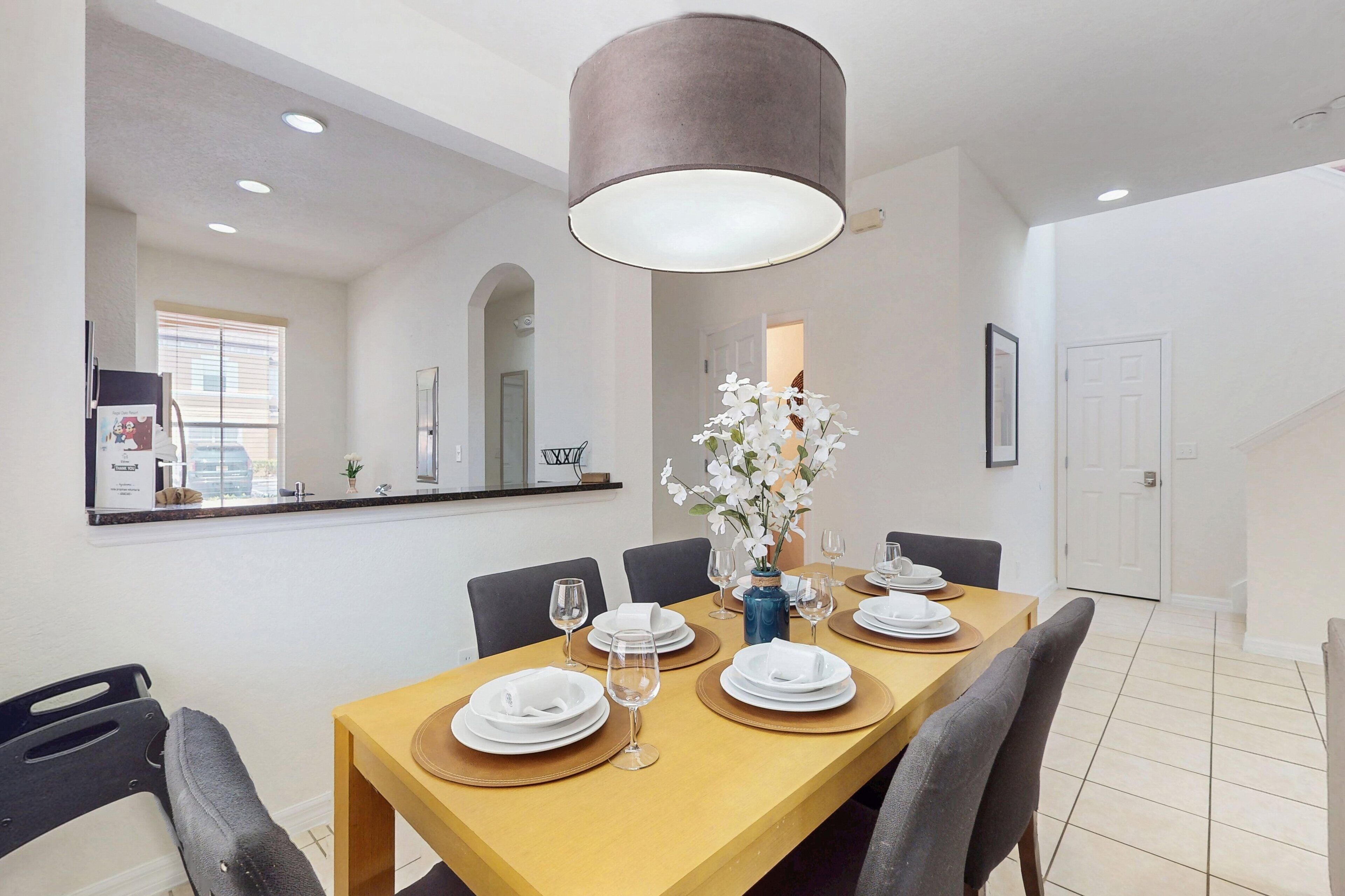 A dining area with a wooden table set for four, a vase with white flowers, and a modern pendant light.