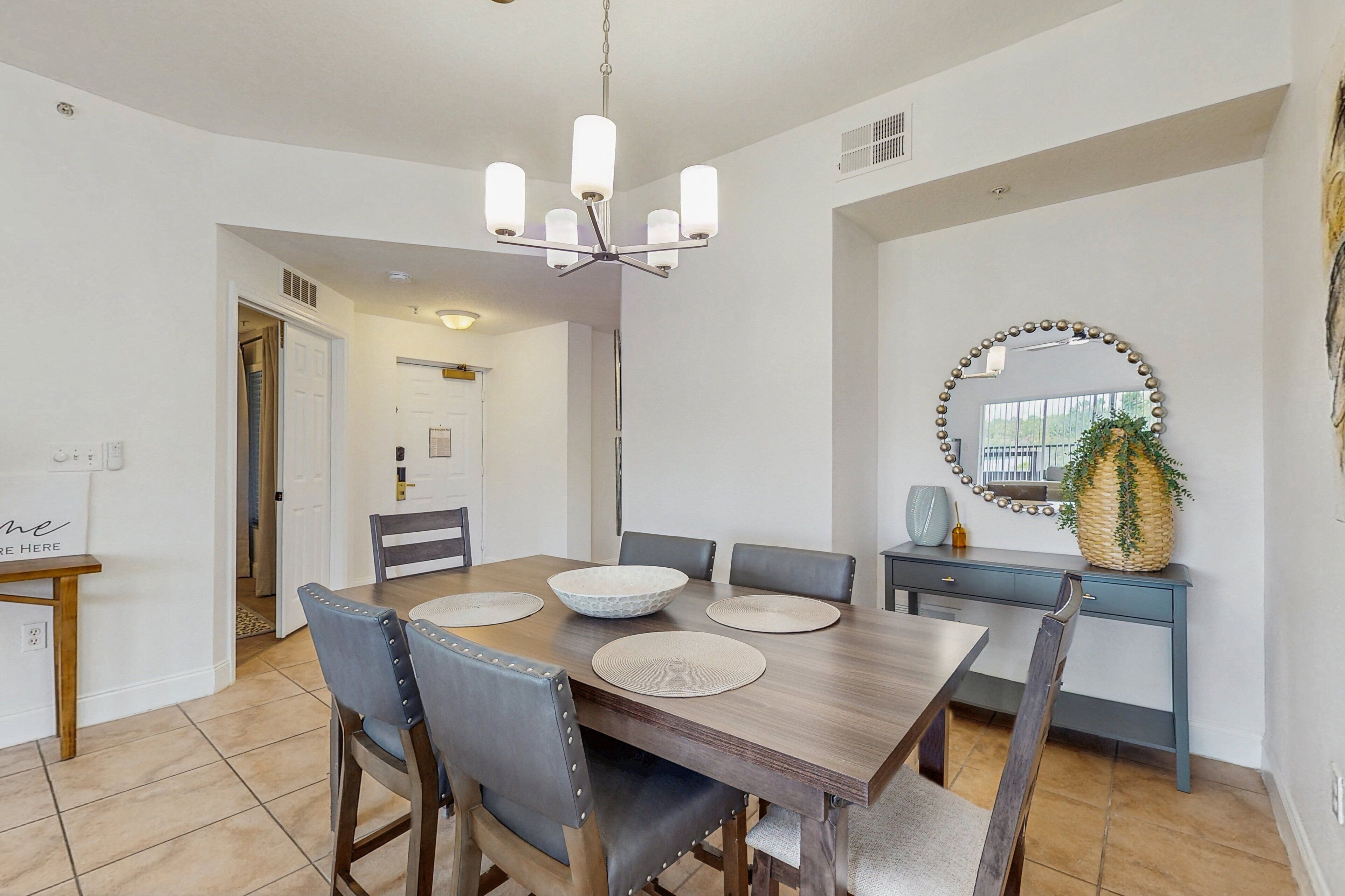 A dining area with a wooden table, six chairs, a chandelier, a mirror, and a plant on a console table.