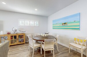 A dining area with a round wooden table, white chairs, a wooden cabinet with books, and a painting of a beach scene.