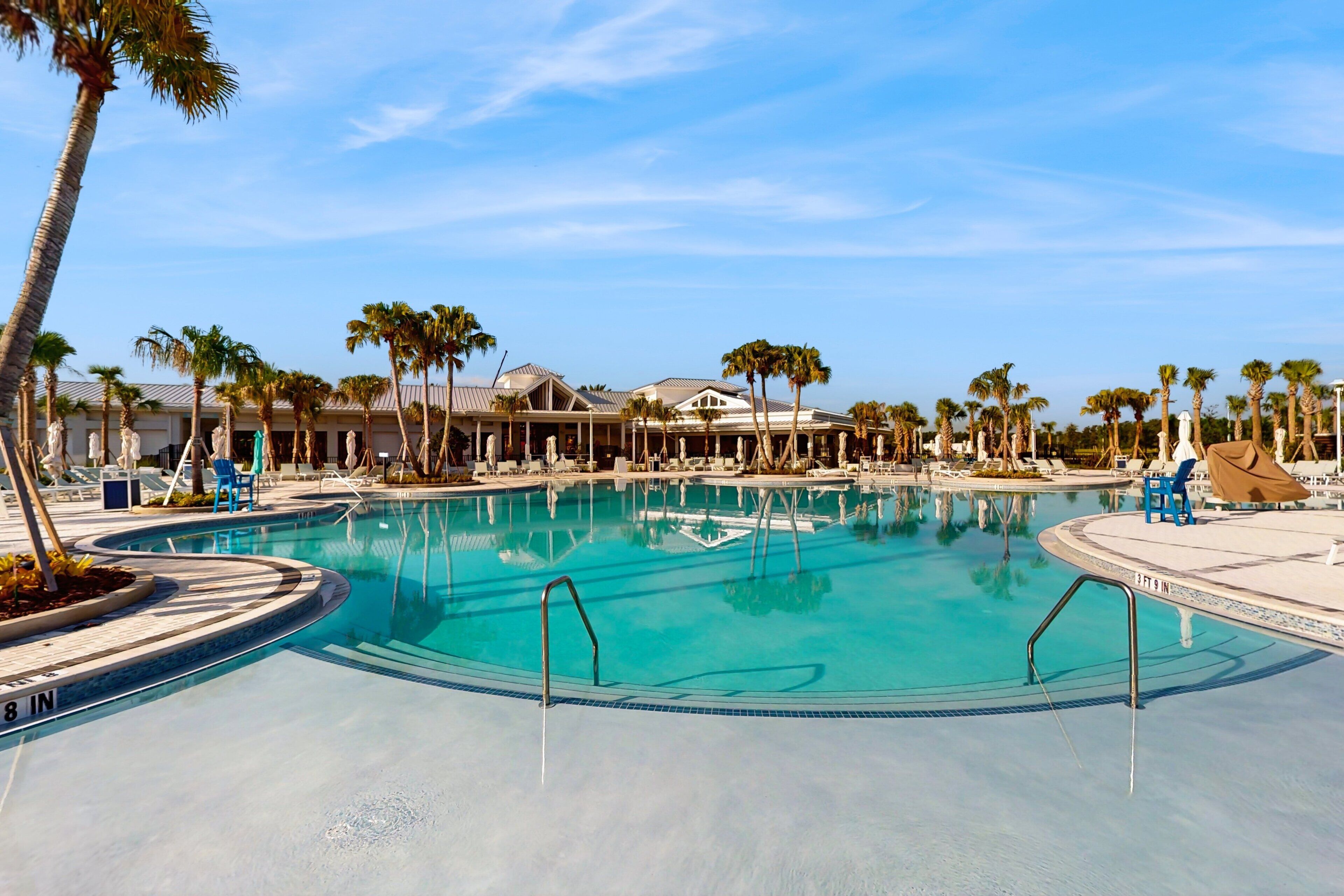 A large outdoor swimming pool with clear blue water, surrounded by palm trees and lounge chairs.